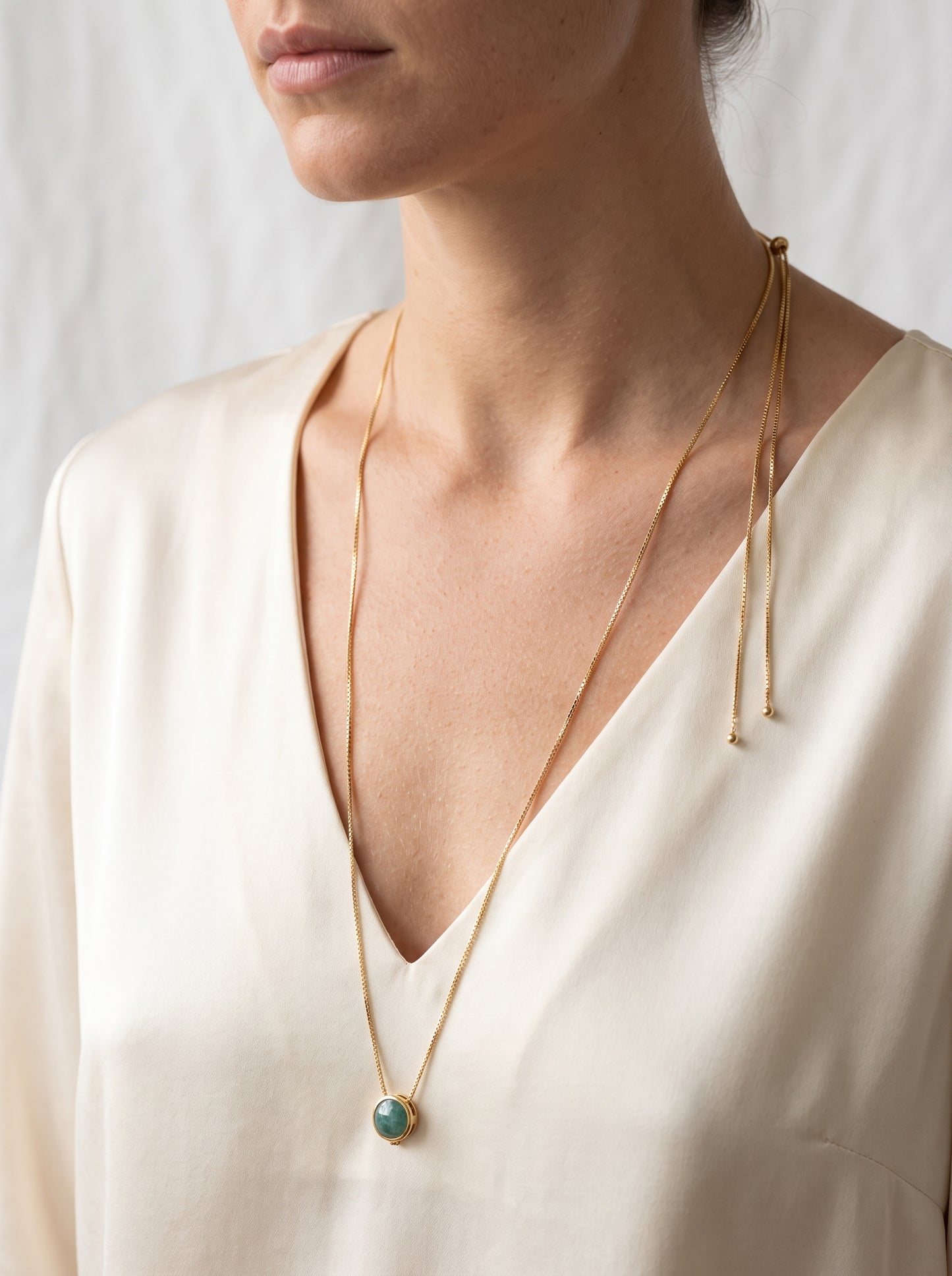 A close-up of a model wearing a delicate gold chain necklace with a small round turquoise pendant, styled with a matching thin gold tie-necklace.
