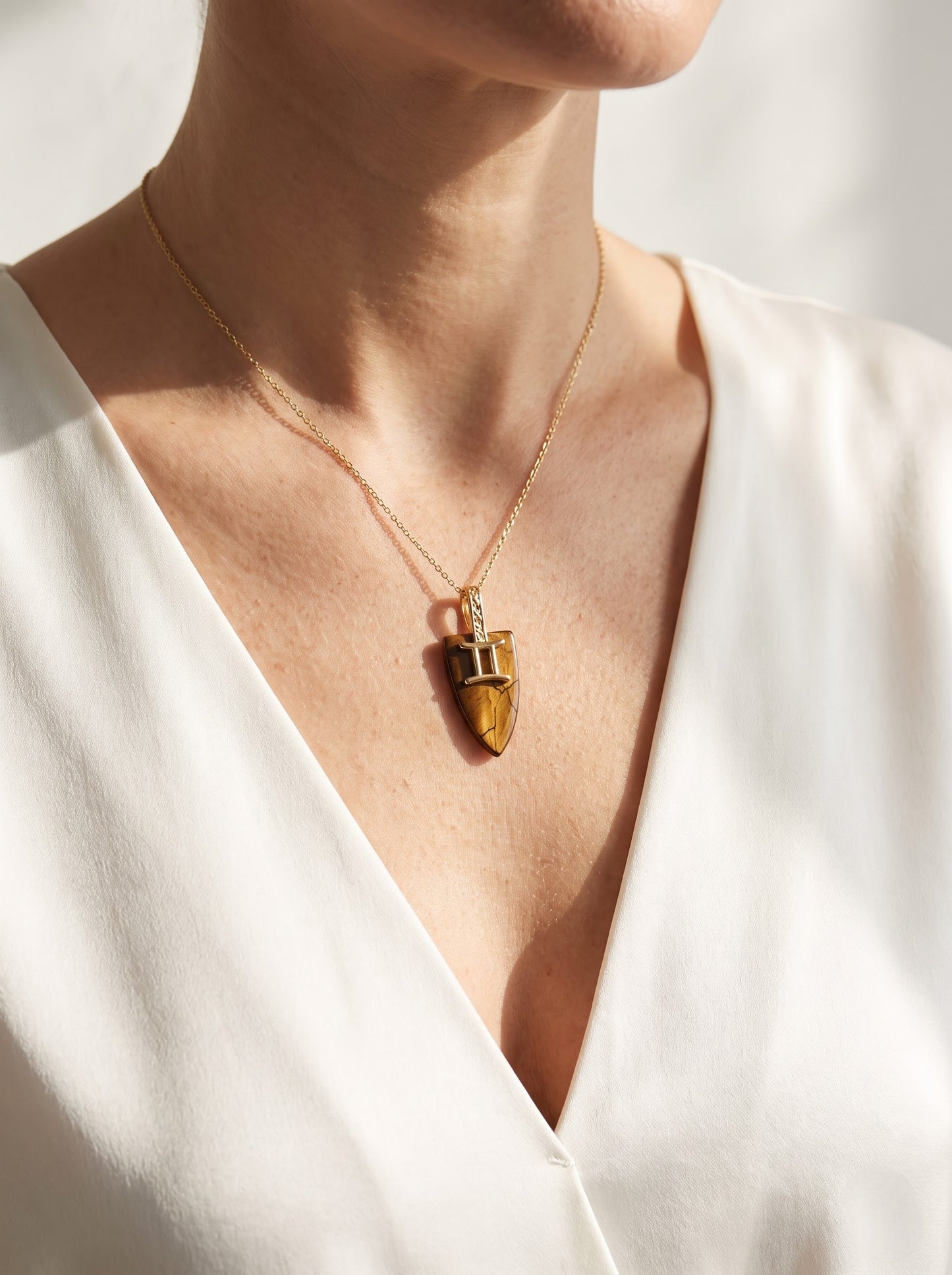 Woman wearing StoneCraft Jewellery tiger's eye Gemini zodiac shield pendant necklace on gold chain, V-neck cream top decolletage shot.