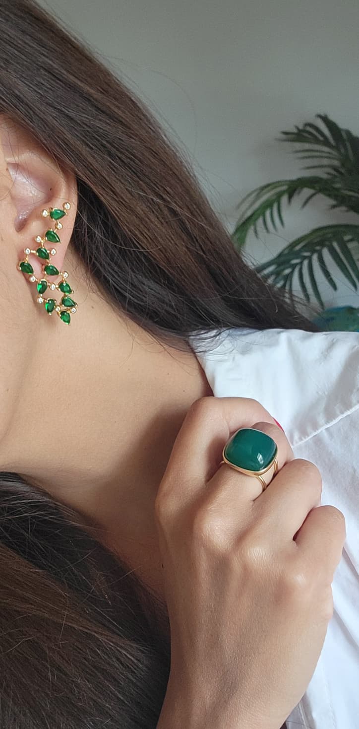 Close-up of a person wearing green gemstone earrings and ring against a neutral background.