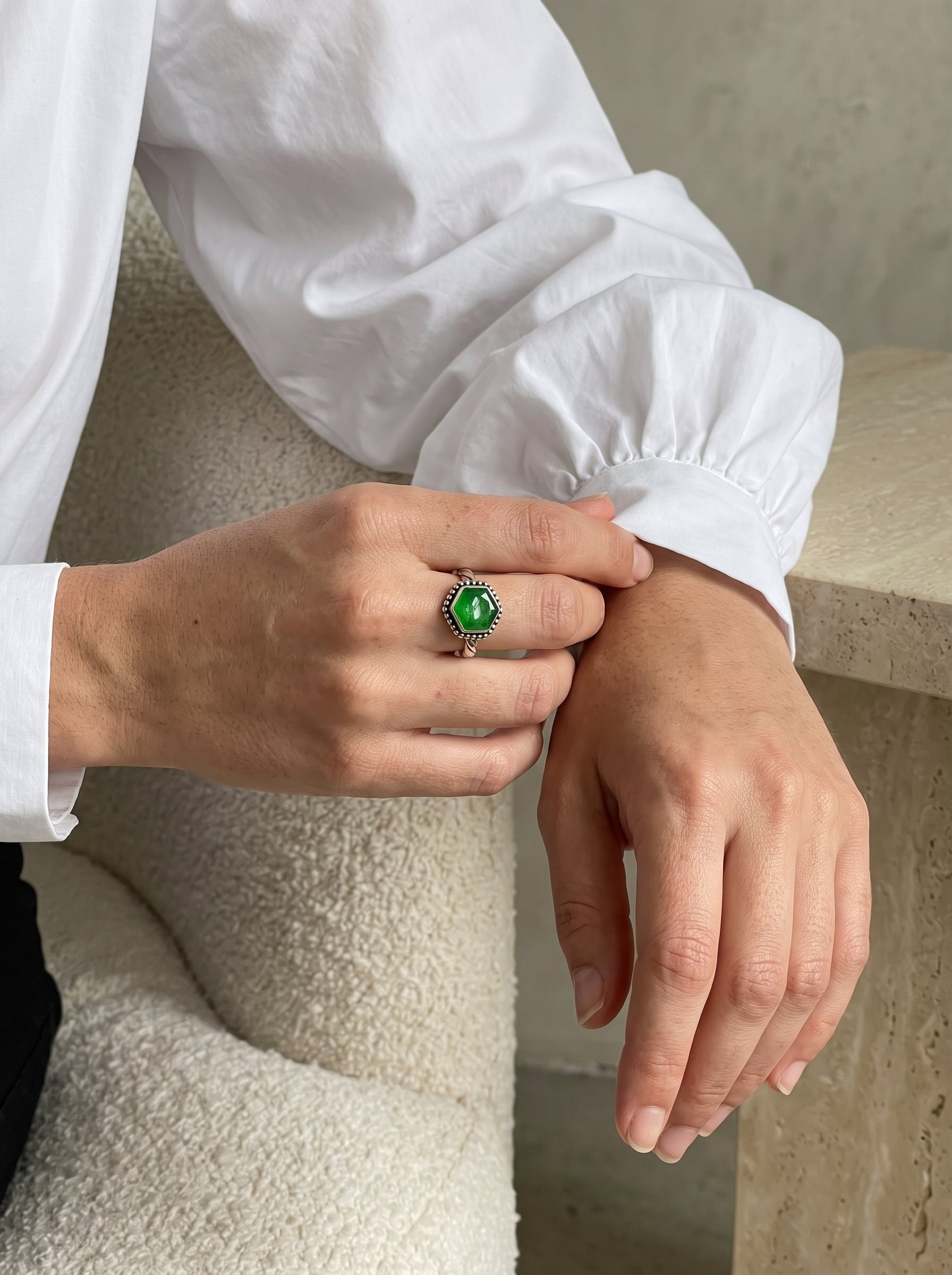 A close-up of a person's hands wearing a silver-toned ring with a green jade gemstone, styled with a white long-sleeve blouse and a textured cream chair.