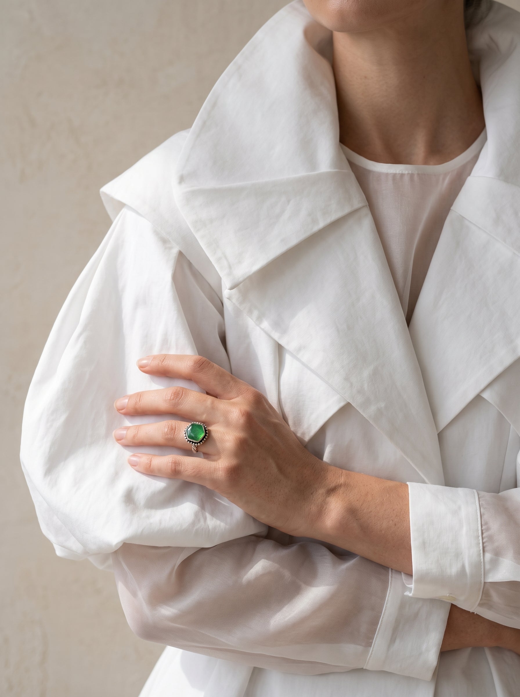 A close-up of a person wearing a white oversized collared shirt or lightweight coat, with one hand resting on the other arm and wearing a ring with a green stone.