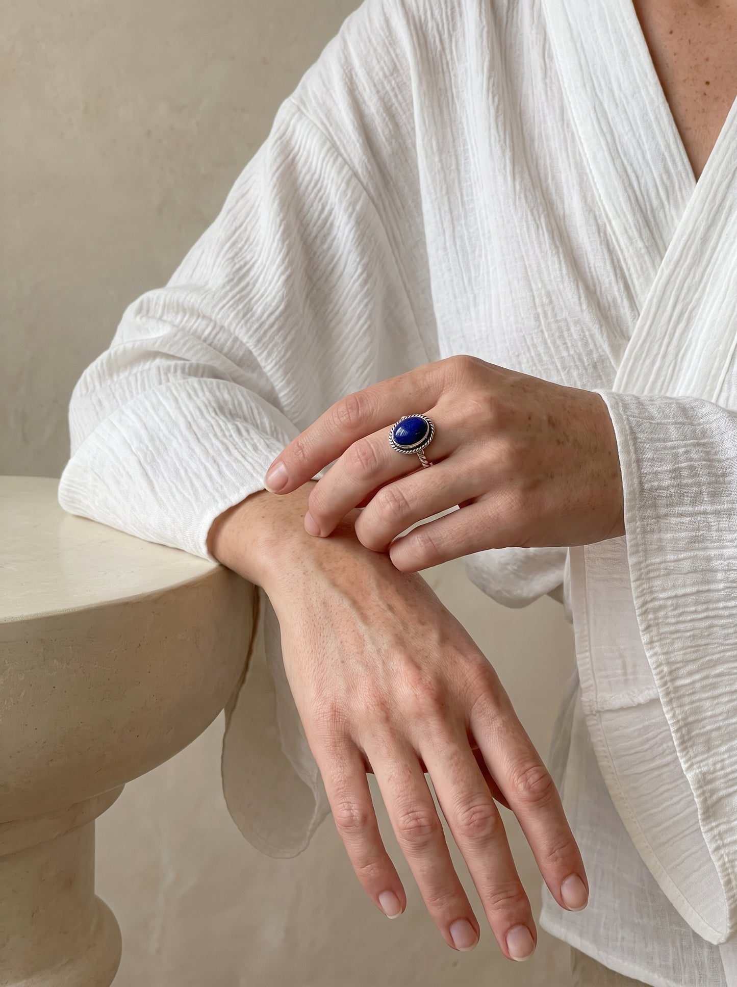 A close-up of a person wearing a silver-toned ring with a deep blue oval stone, resting one hand on the other. The person is wearing a textured white long-sleeve blouse and leaning on a light beige stone pedestal.