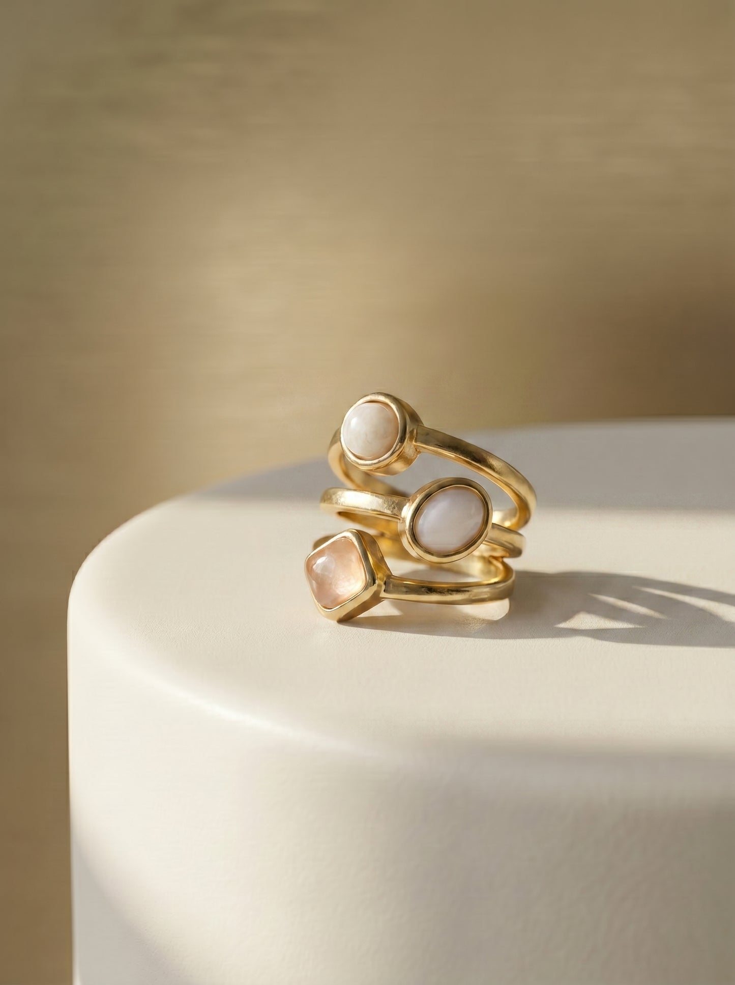 Gold multi-stone ring with mother of pearl and rose quartz stones displayed on a white curved surface against a warm beige background