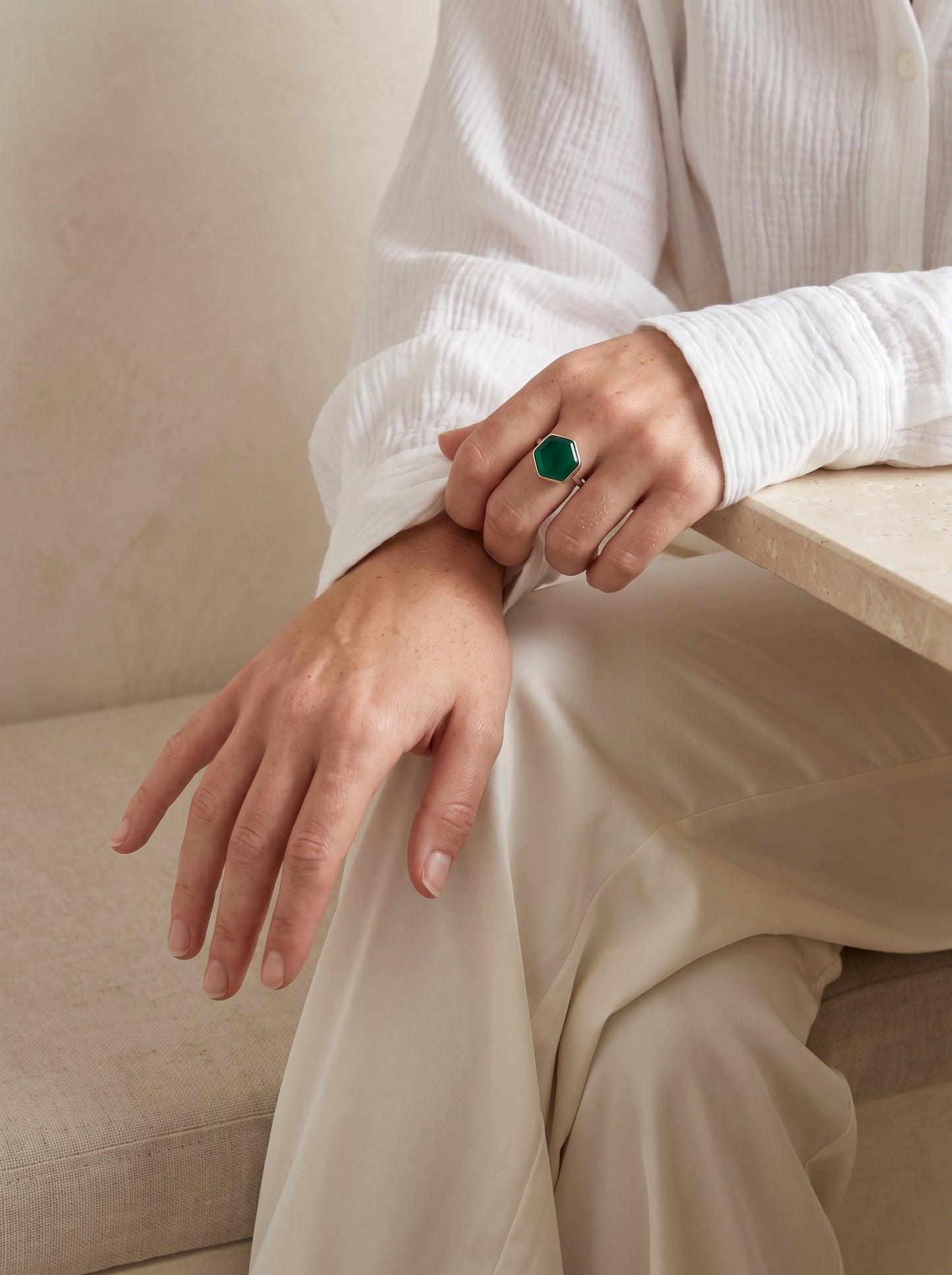 A close-up of a person wearing a silver ring with a green marquise-cut green onyx gemstone, styled with a white textured blouse and beige trousers while sitting on a beige sofa.