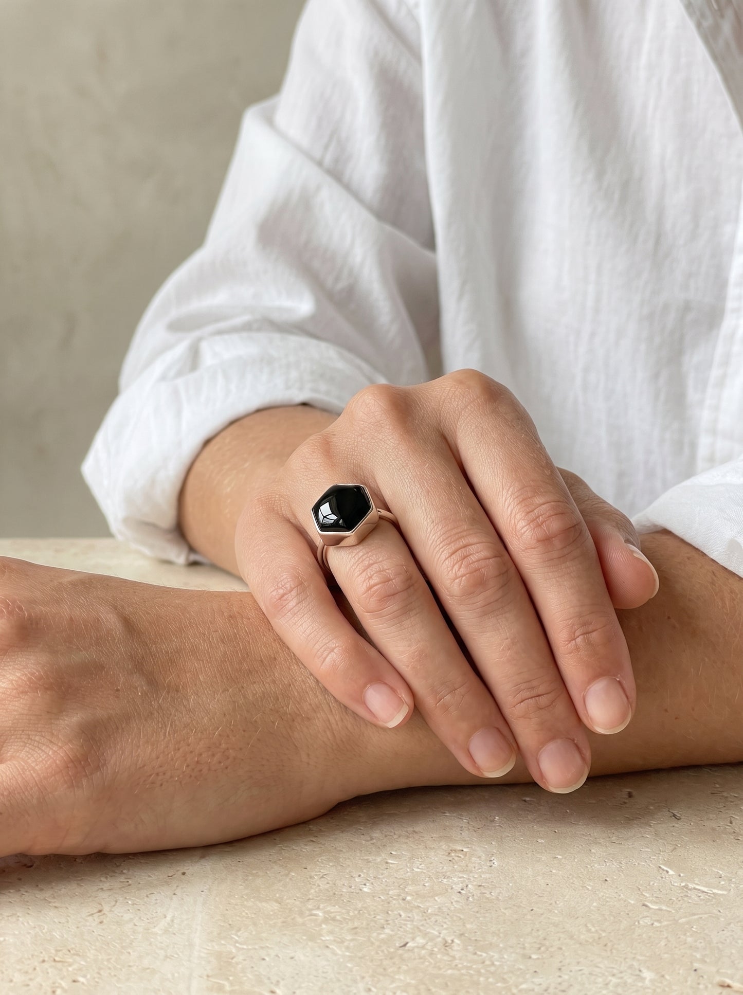 A close-up of a person's hand wearing a silver ring with a faceted black  onyx gemstone, resting on their forearm against a neutral beige background.