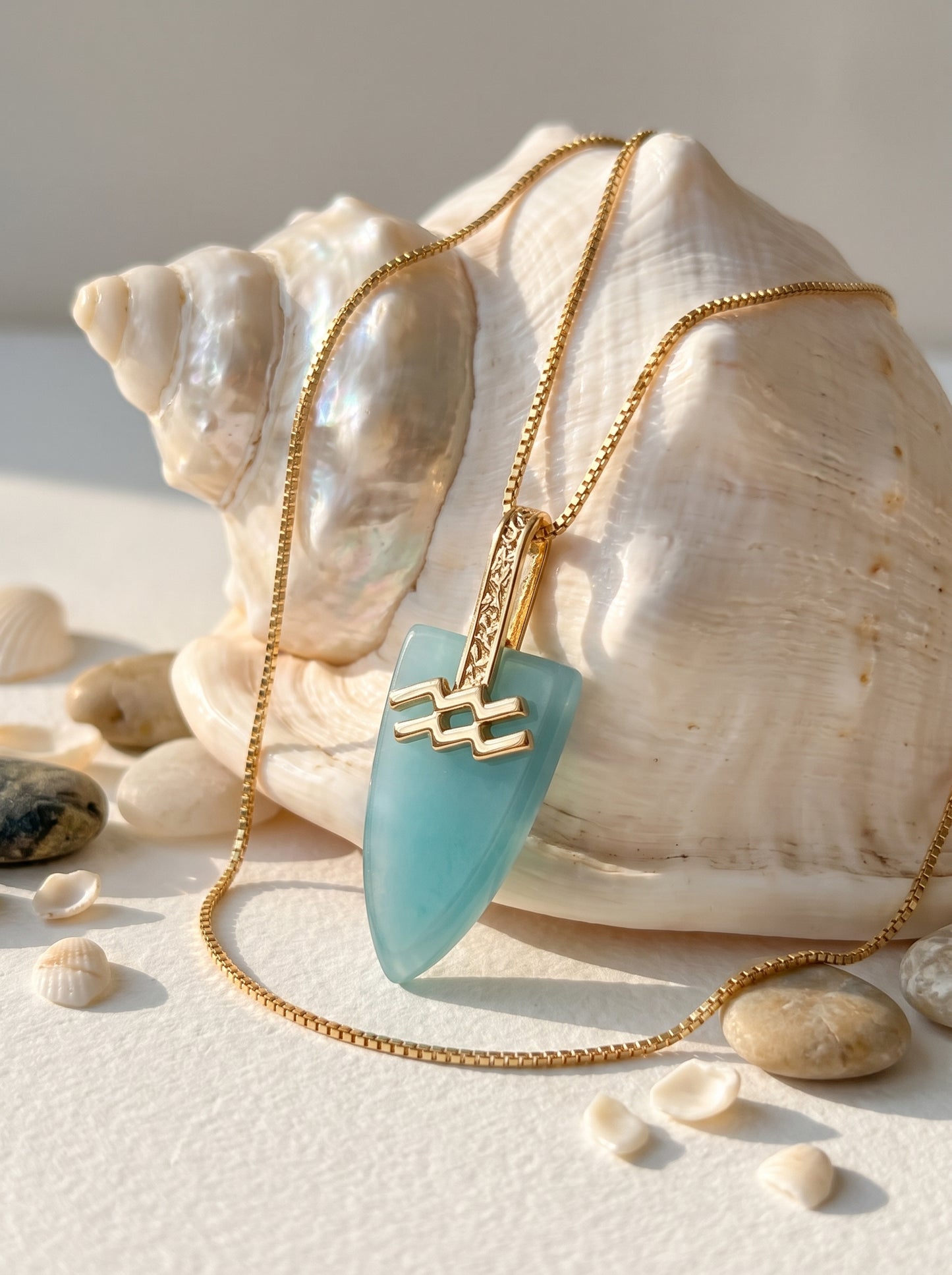 : Aquarius agate pendant necklace with gold-plated box chain laid against a large white shell surrounded by pebbles and small shells