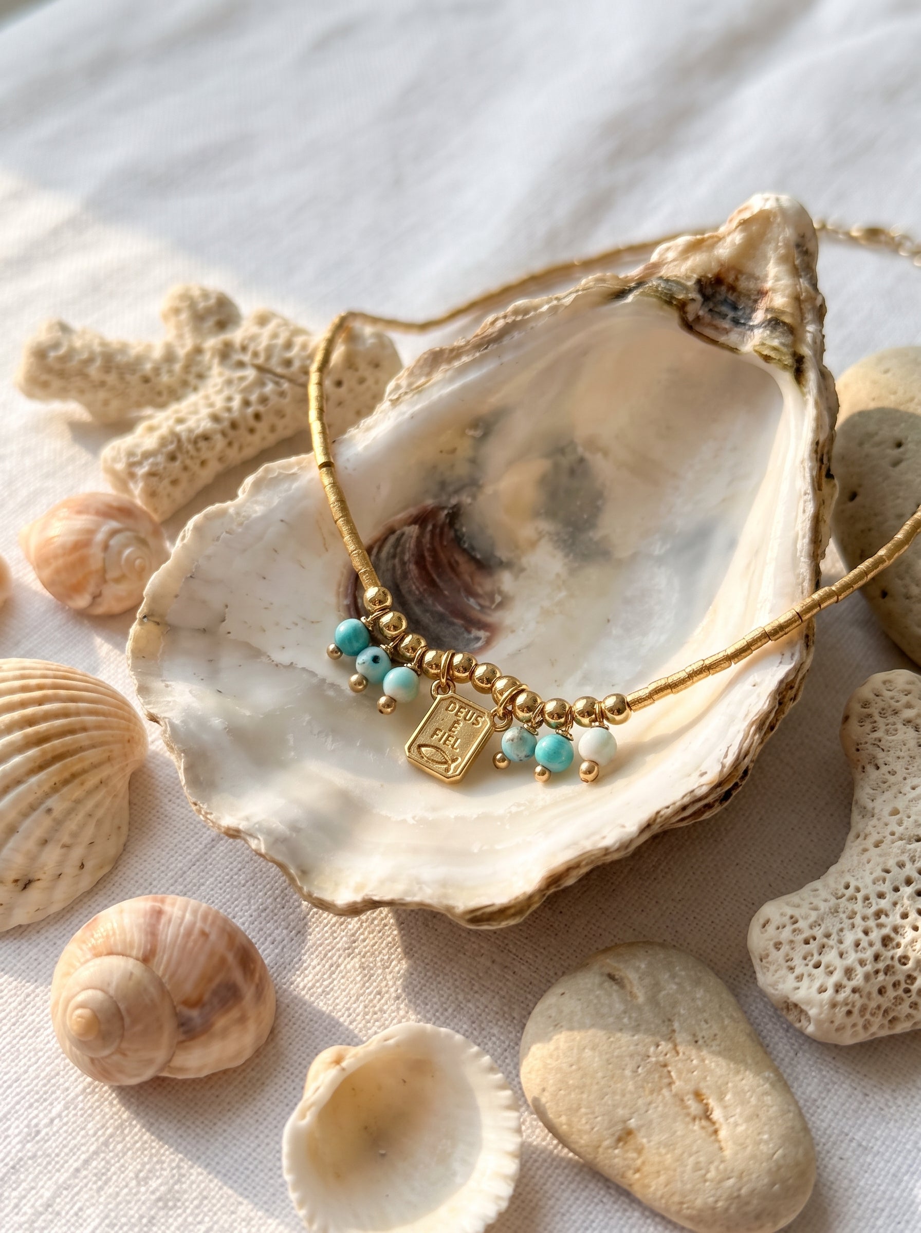 A delicate gold necklace with turquoise beads and a small gold charm, displayed inside an open seashell surrounded by natural shells and coral on a light fabric surface.