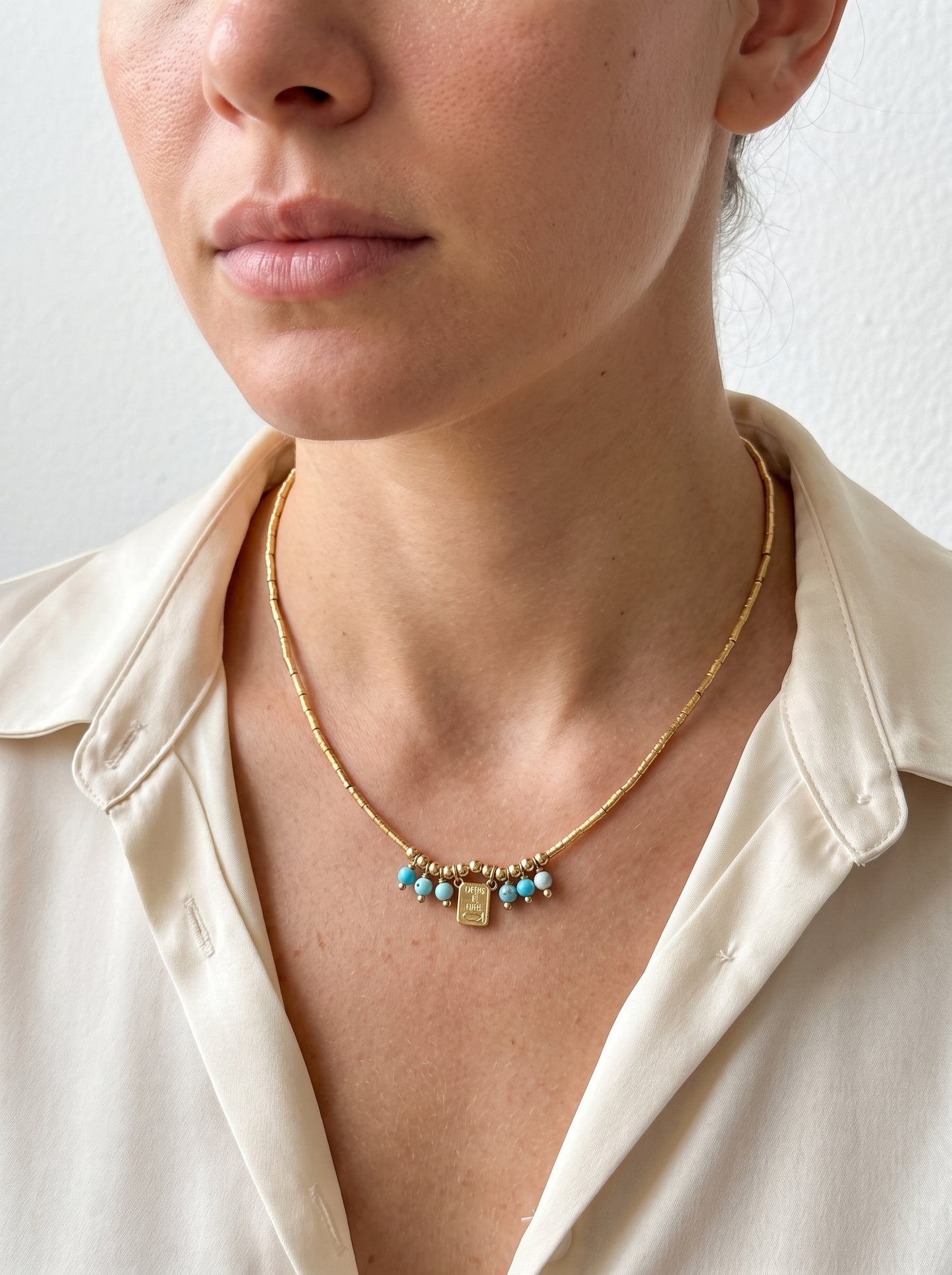 A close-up of a model wearing a delicate gold chain necklace with small turquoise blue beads and a central gold charm, paired with a cream-colored satin shirt.