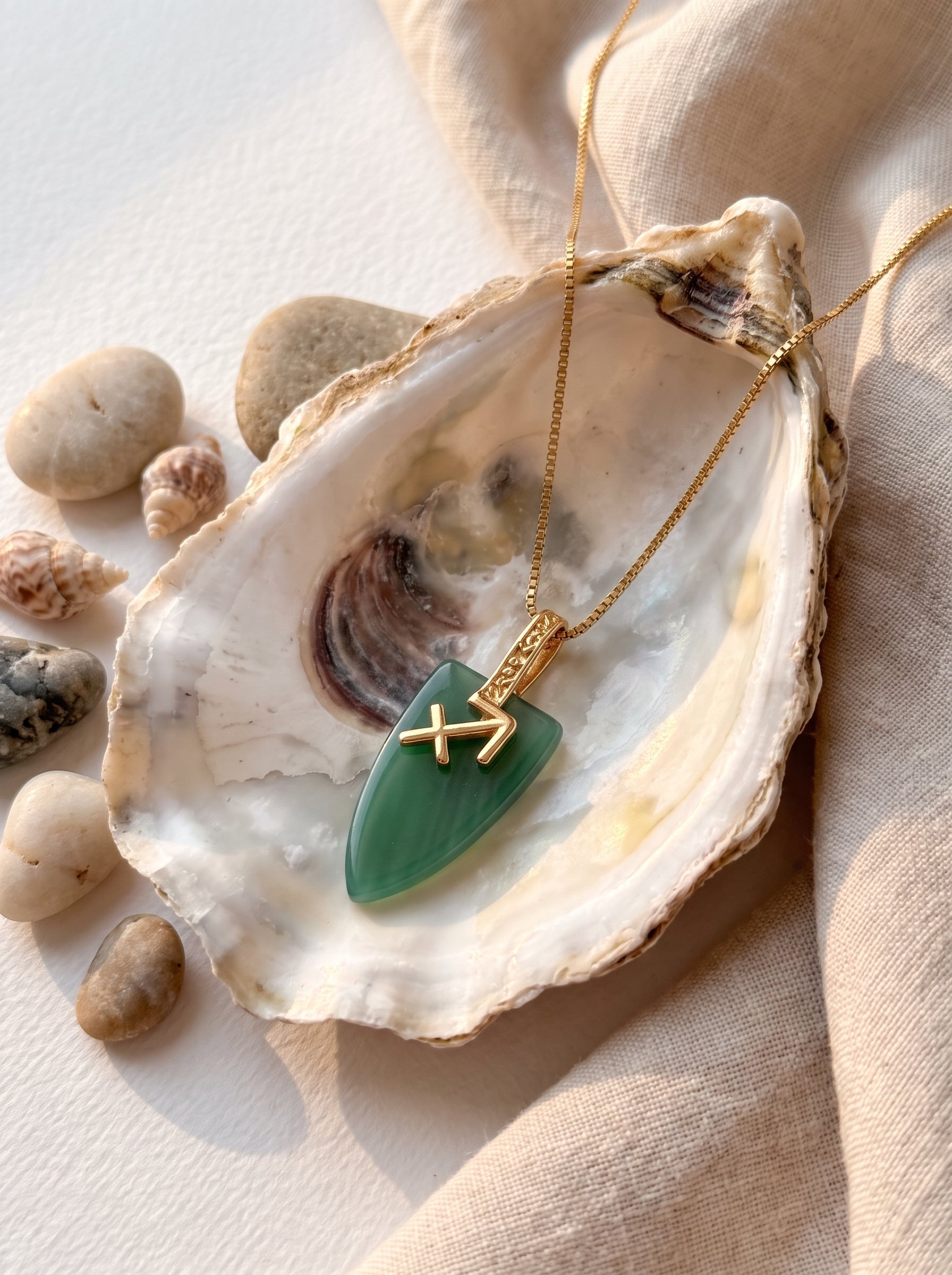 Sagittarius agate arrowhead pendant necklace with gold zodiac symbol resting inside an oyster shell surrounded by pebbles and small shells
