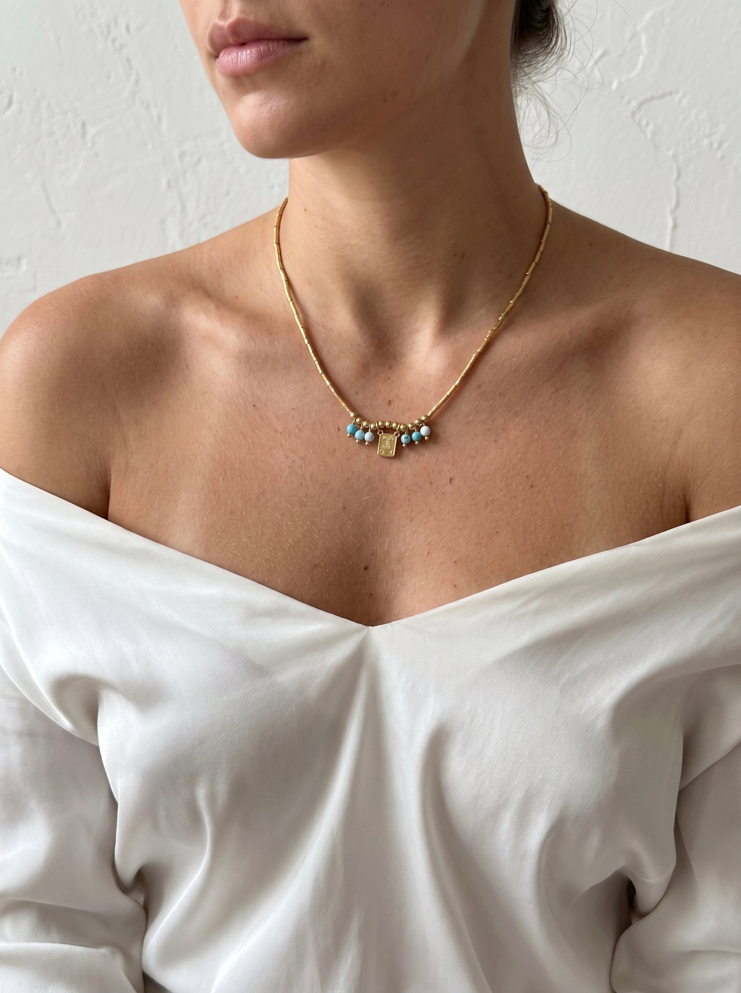 A close-up of a model wearing a delicate gold chain necklace with small blue and gold beads at the center, paired with an off-the-shoulder white top against a textured white wall.