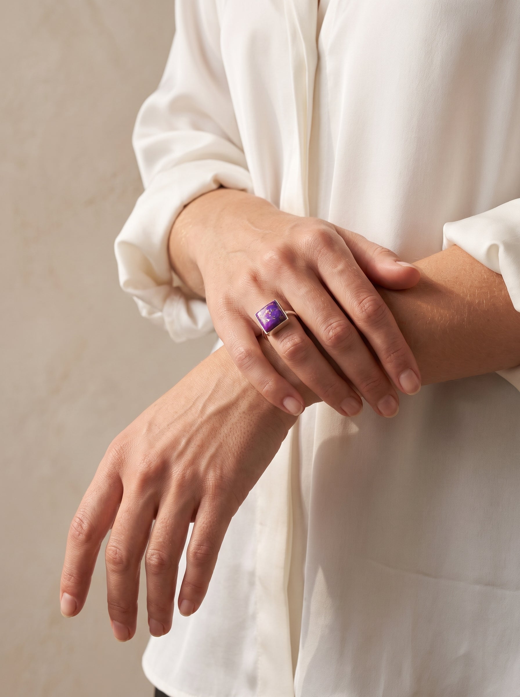 A close-up of a person wearing a gold ring with a square purple stone, shown on their middle finger while wearing a white blouse against a neutral beige background.