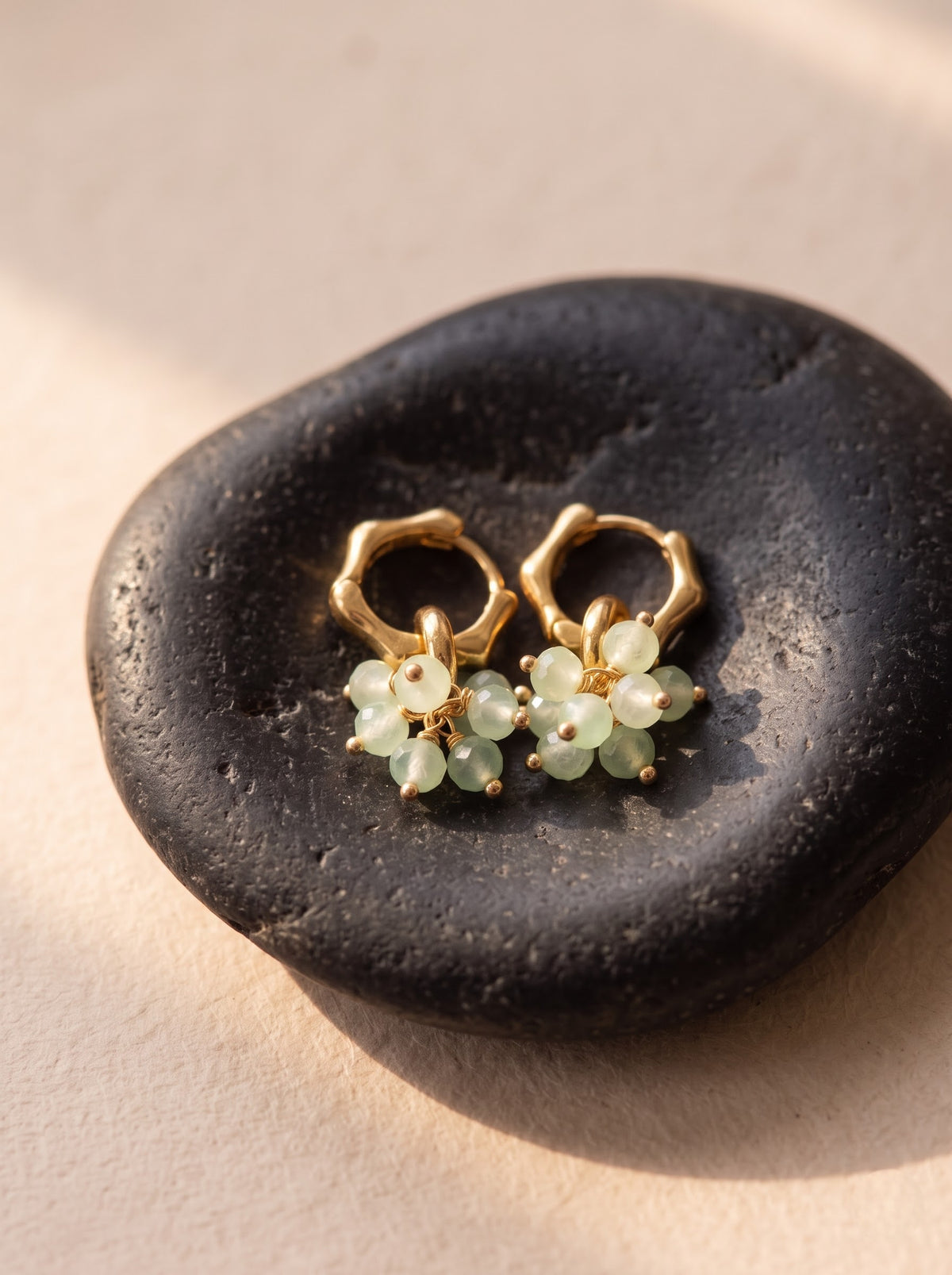 A pair of gold hoop earrings with small pale green aventurine clusters, displayed on a dark smooth stone against a warm beige background.