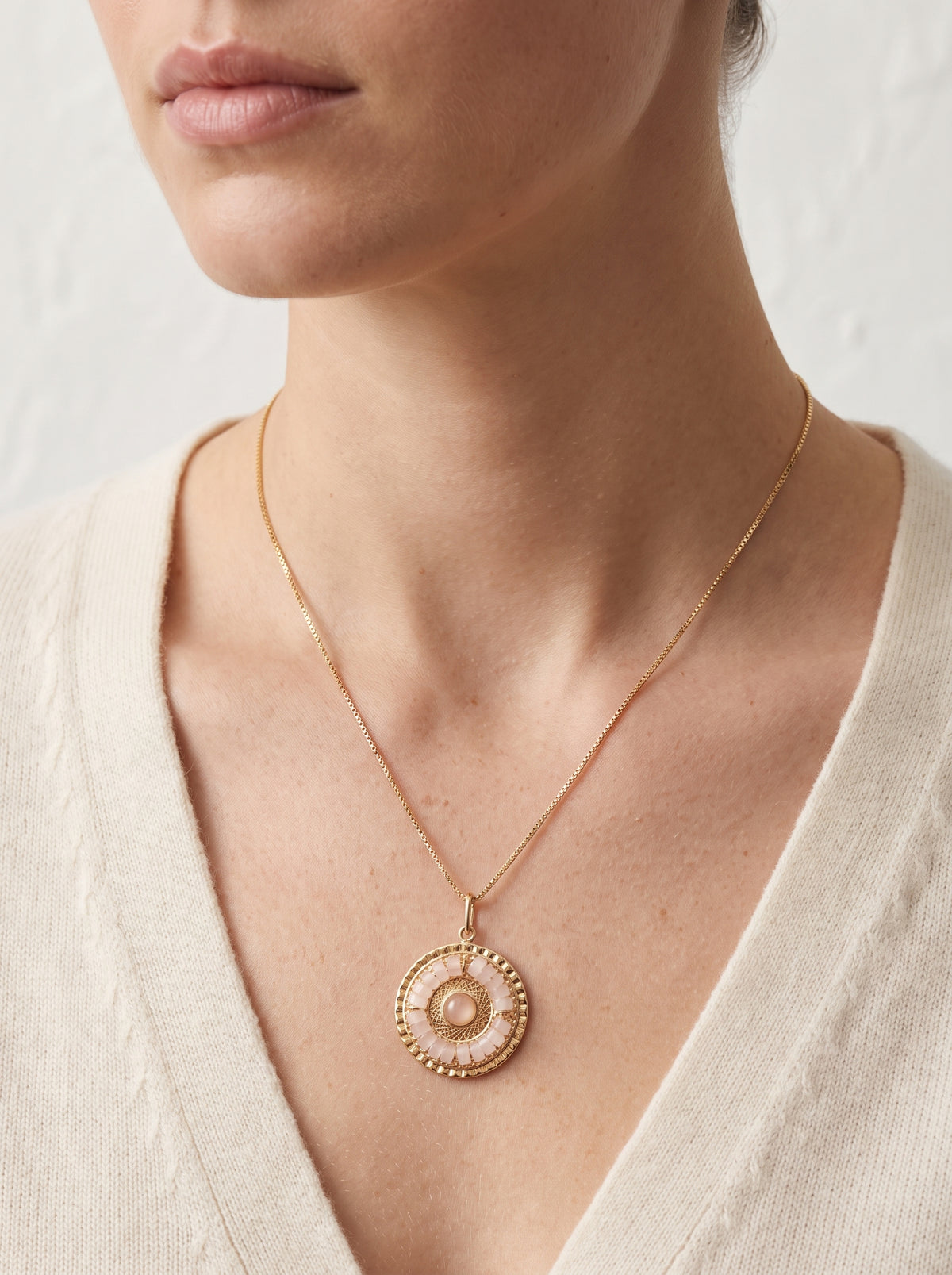 A close-up of a model wearing a delicate gold chain necklace with a mandala rose quartz  pendant featuring a textured beaded border and a central pearl-like accent, paired with a cream-colored knit top.