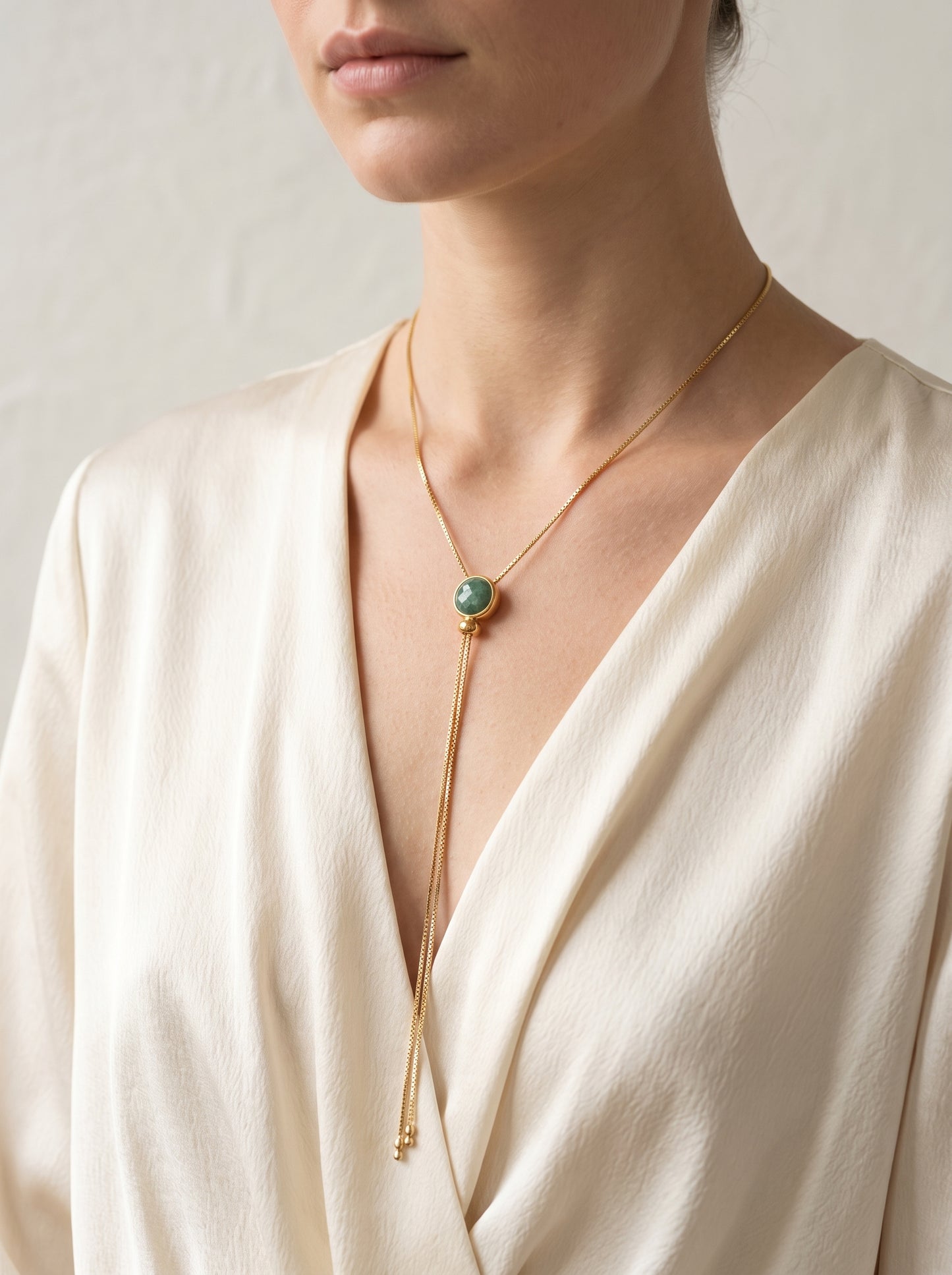 A close-up of a model wearing a delicate gold chain necklace with a round green stone pendant and long dangling chains, styled with a cream-colored wrap top.