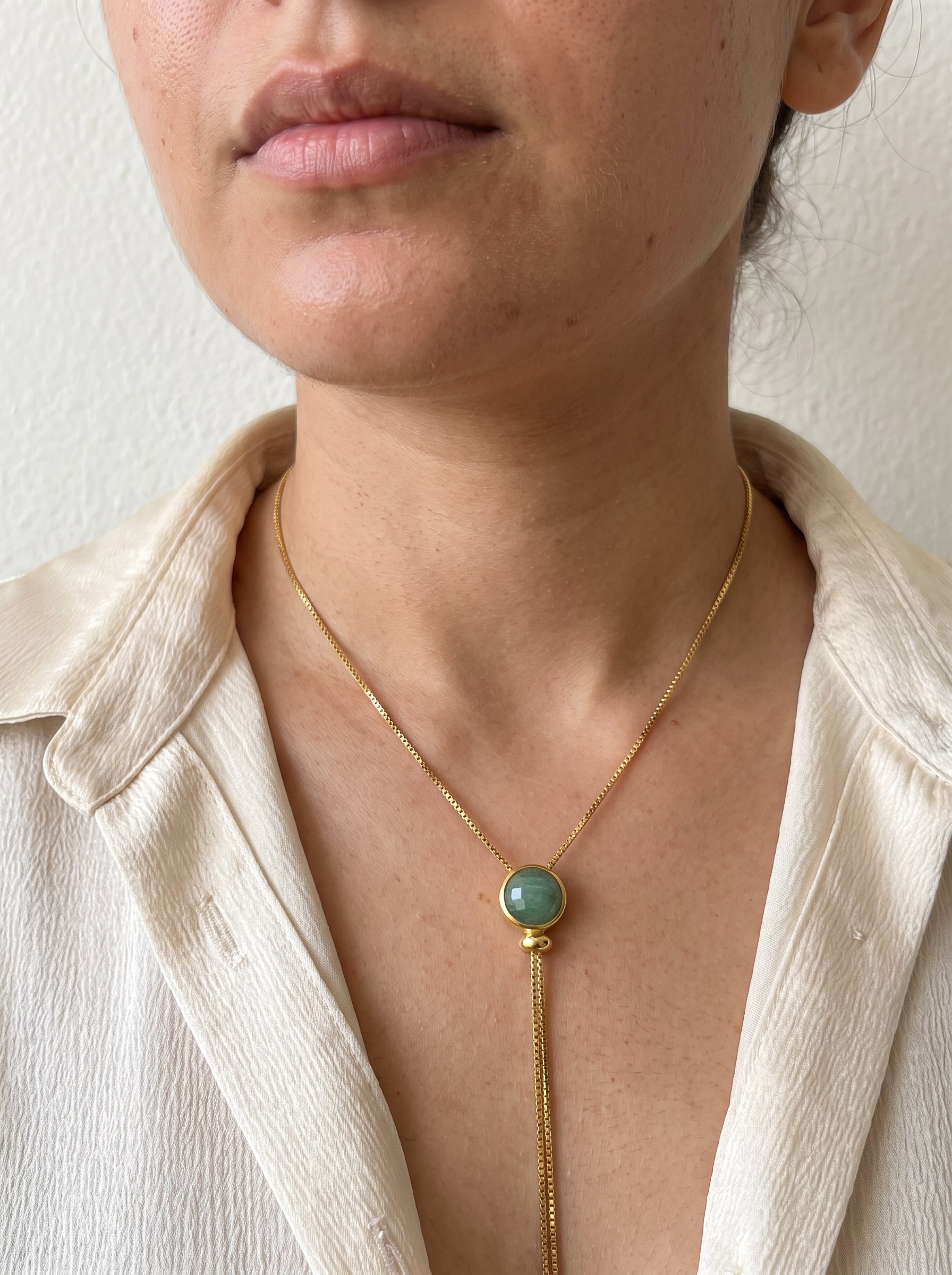 A close-up of a model wearing a delicate gold chain necklace with a round light green stone pendant and a long drop chain.