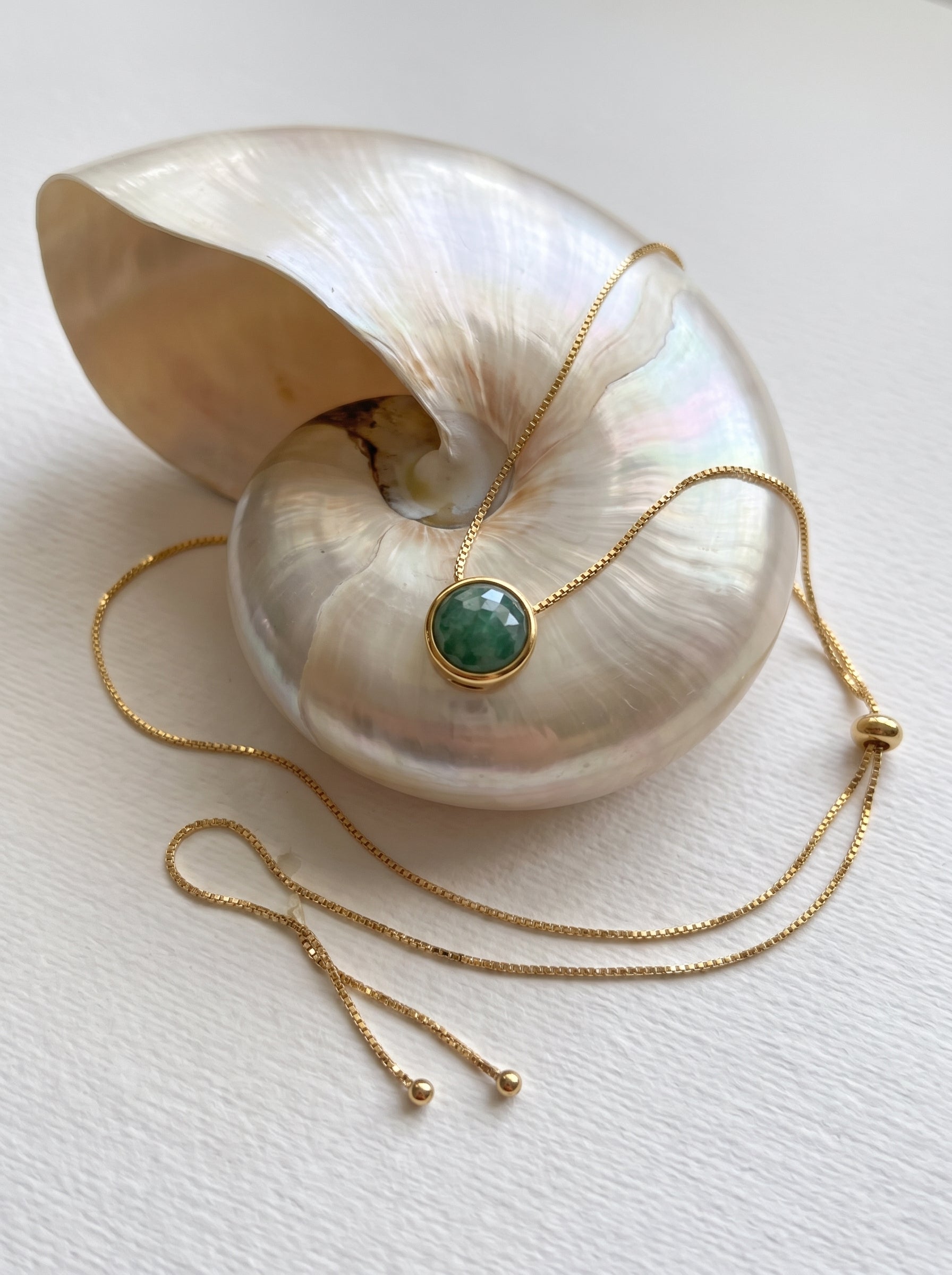 A gold chain lariat  necklace with a round green  quartz gemstone pendant, draped over a large iridescent shell on a light textured background.
