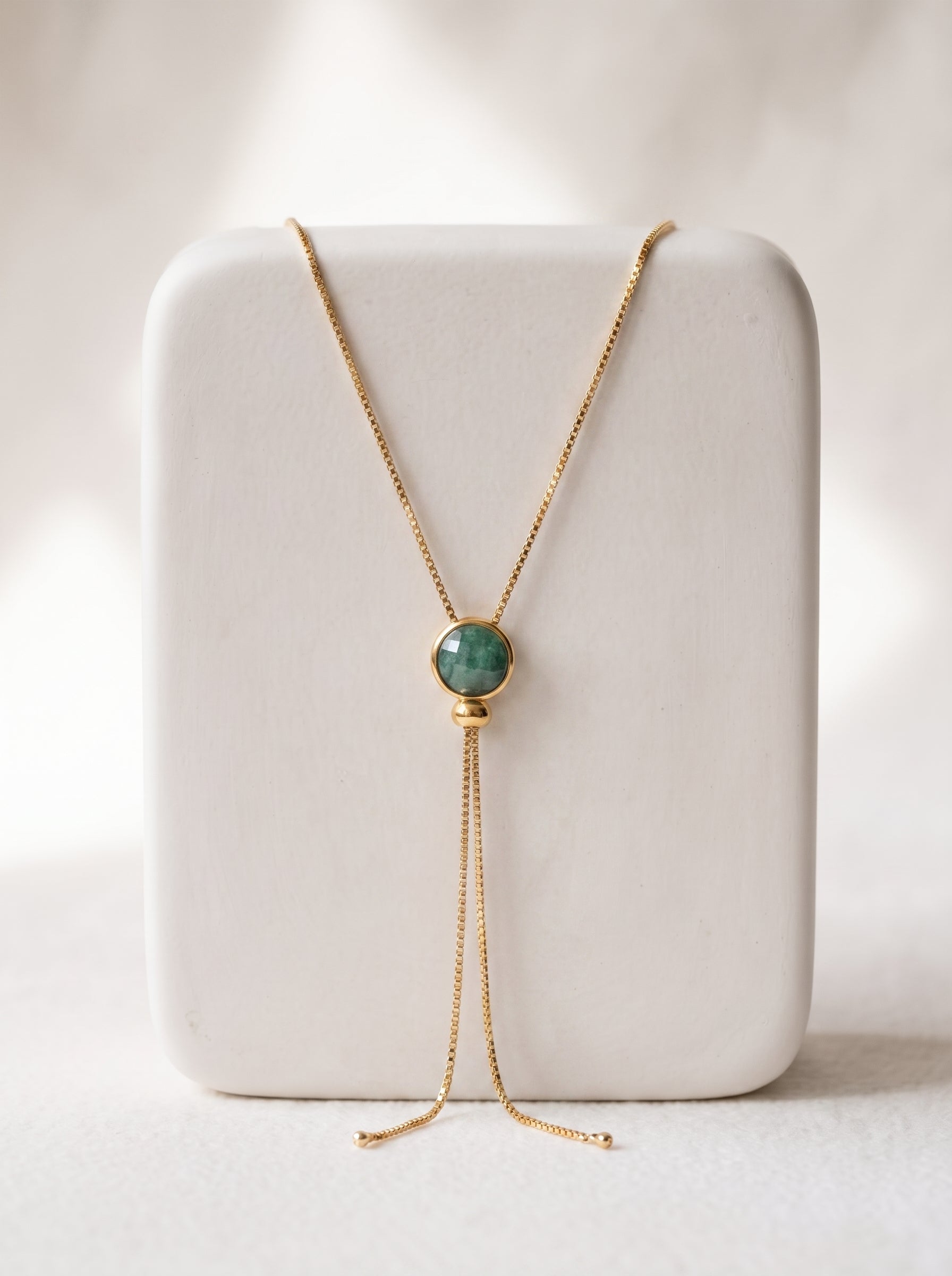 A gold plated necklace with a round green quartz stone pendant, displayed on a white jewelry stand against a soft, light background.