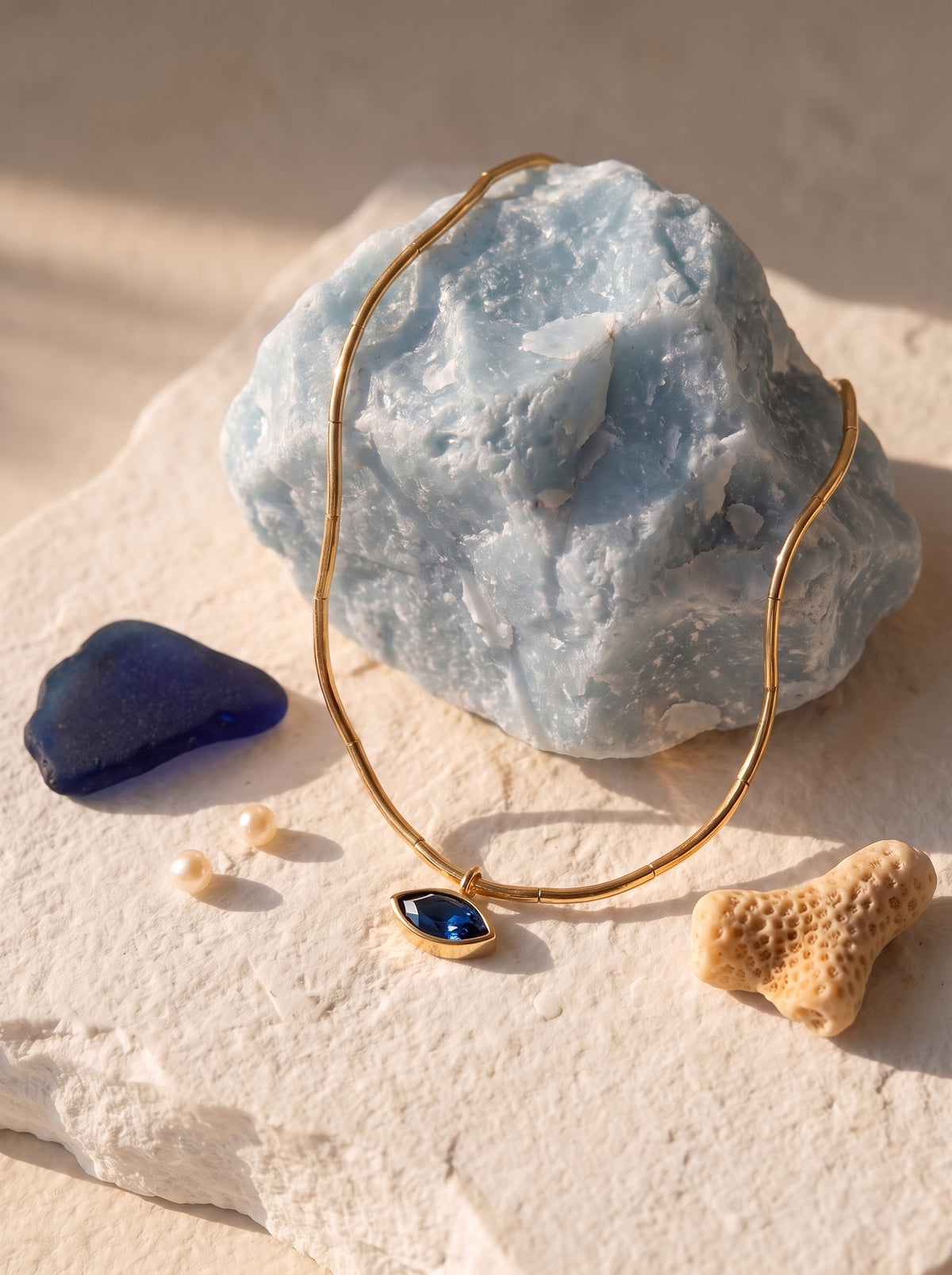  StoneCraft Jewellery Gold Wave Chain Necklace with Blue Crystal Pendant displayed on blue stone rock with sea glass and coral