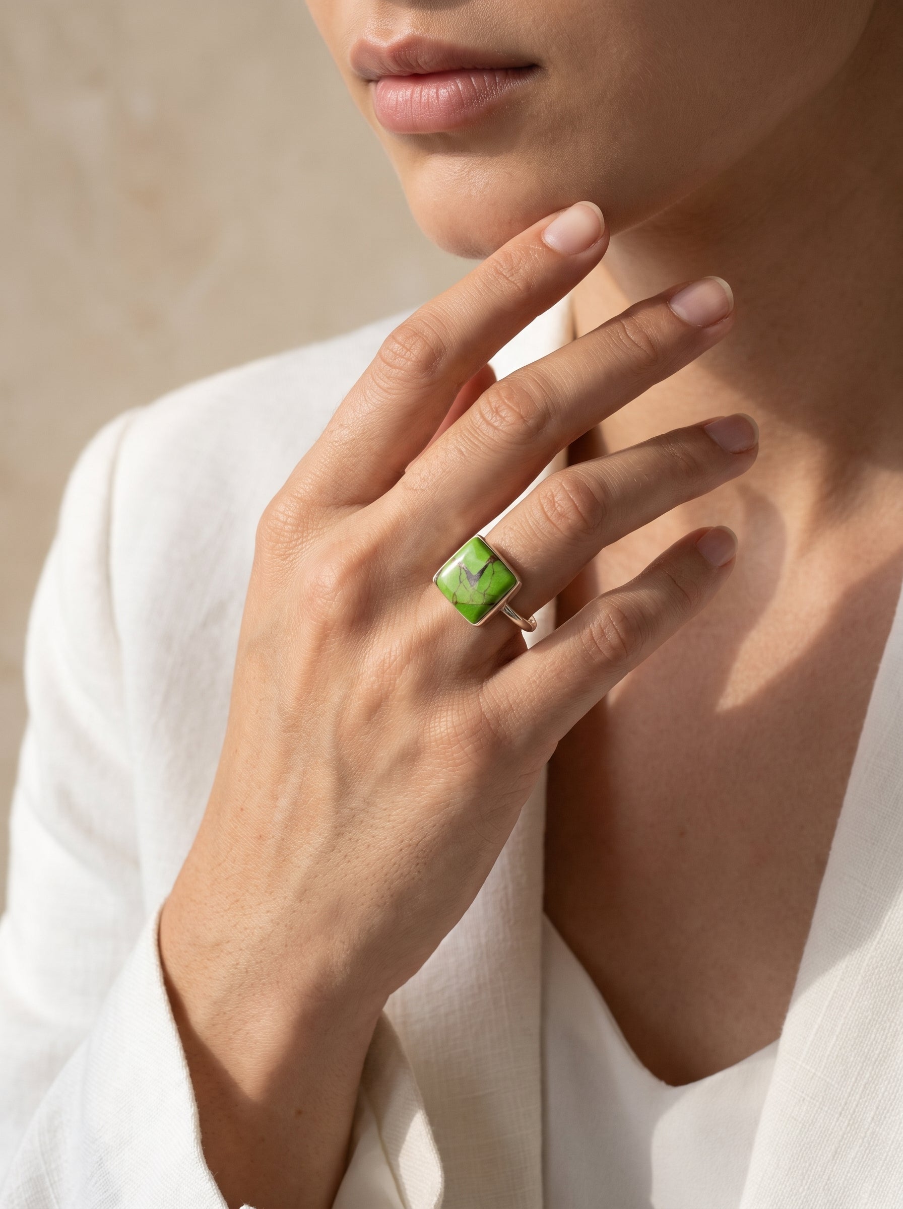 A close-up lifestyle shot of a model wearing a gold ring with a square green stone, set against a soft beige background and white blazer.