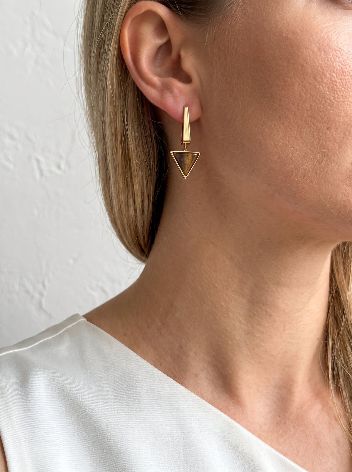 Close-up of a model wearing StoneCraft Jewellery gold-plated Tiger’s Eye triangle drop earrings with a white top.