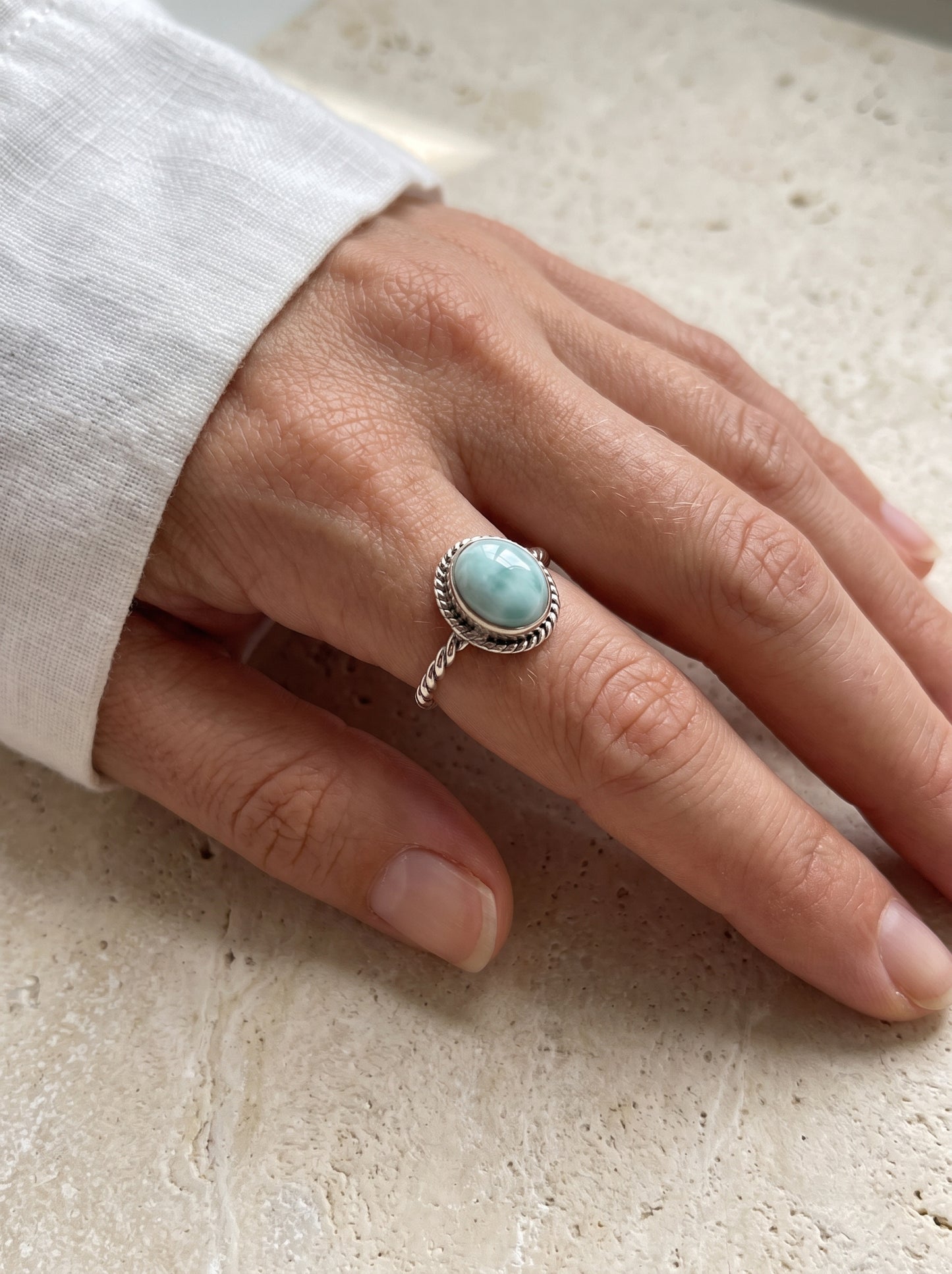 A close-up of a hand wearing a silver ring with a round light blue-green stone, resting on a textured beige stone surface.