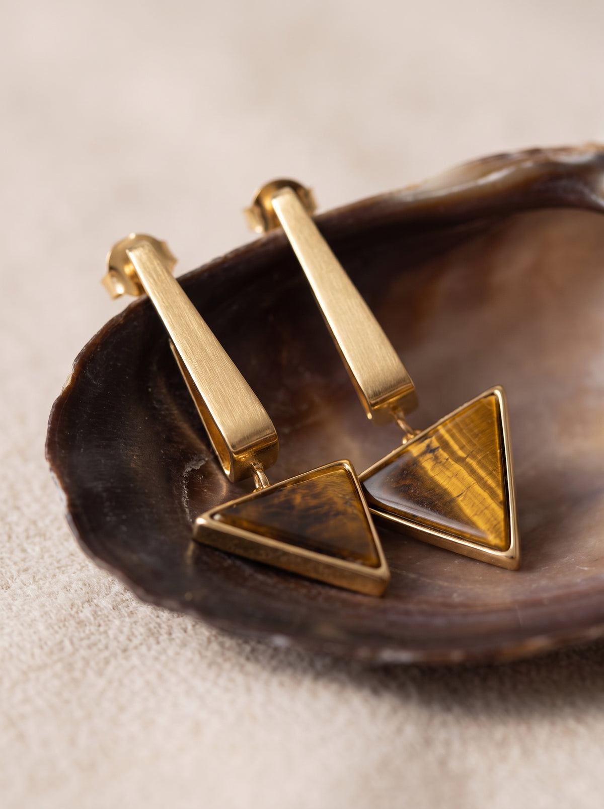 Pair of StoneCraft Jewellery gold-plated Tiger’s Eye triangle drop earrings on a round white display.