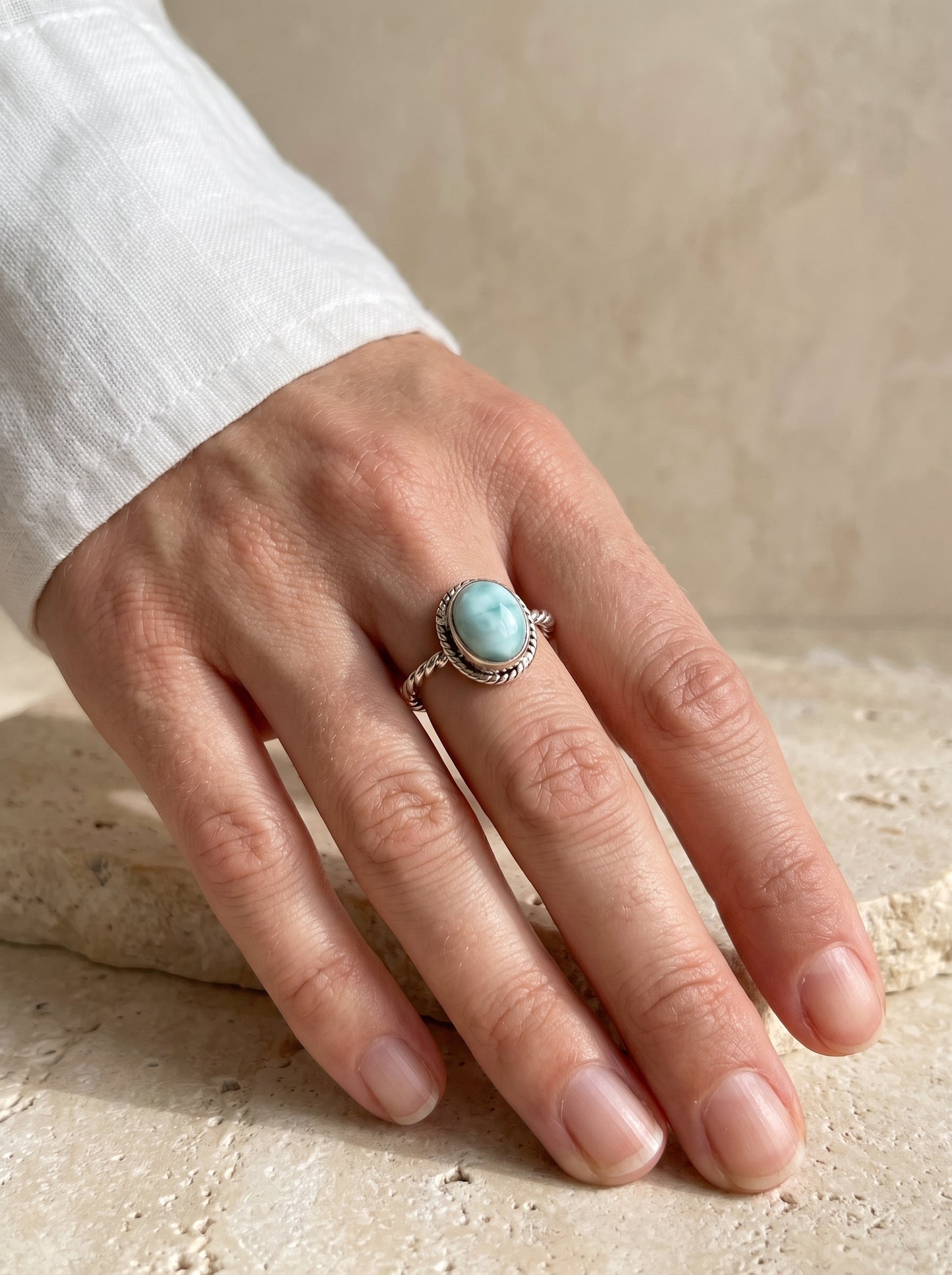 A silver ring with a round larimar worn on a person's hand against a neutral beige stone background