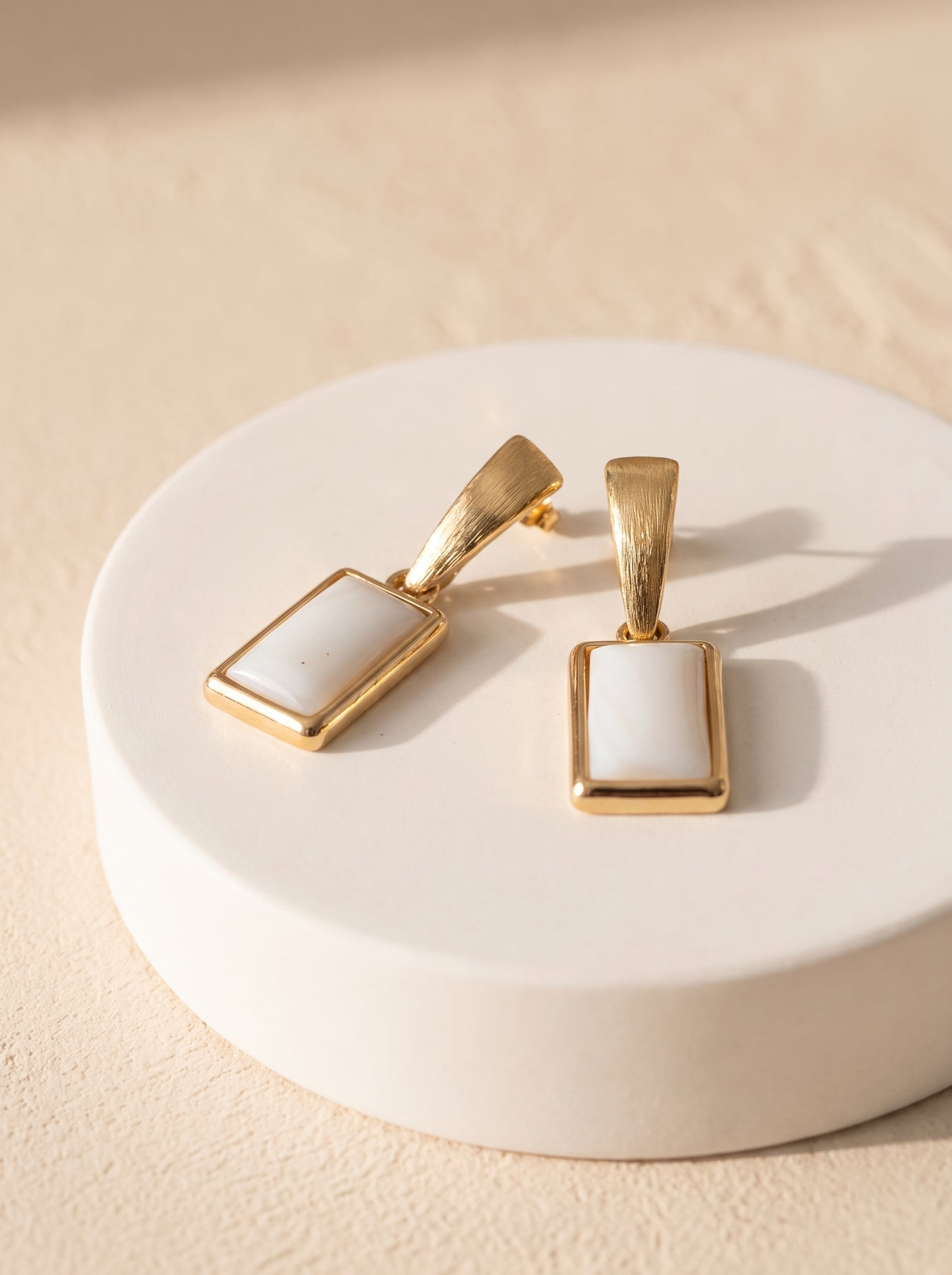 A pair of gold rectangular pendant earrings with mother of pea, displayed on a round white pedestal against a textured beige background.l