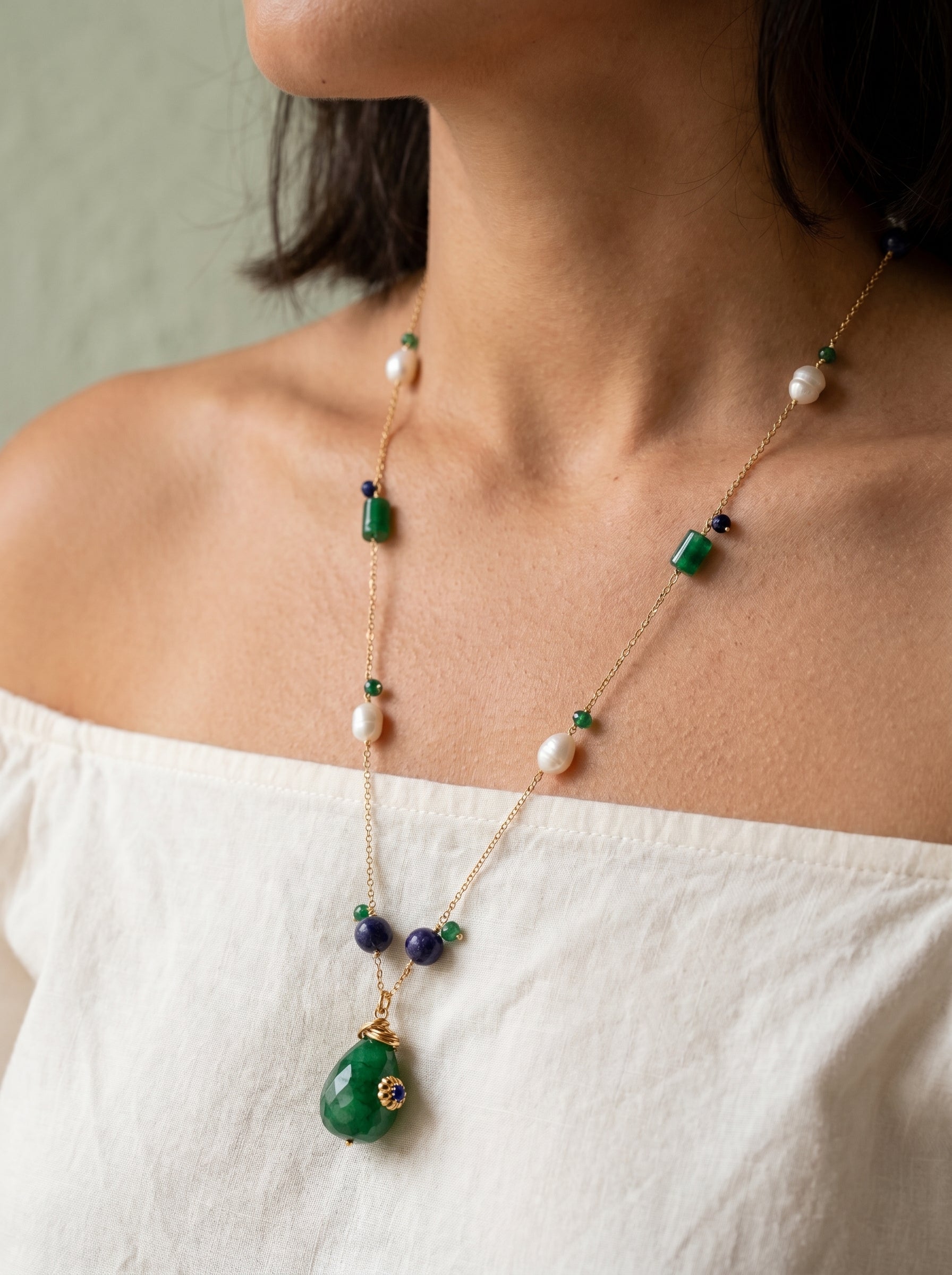 Woman wearing gold-plated green jade and pearl long necklace with sapphire beads against a sage green background, white top