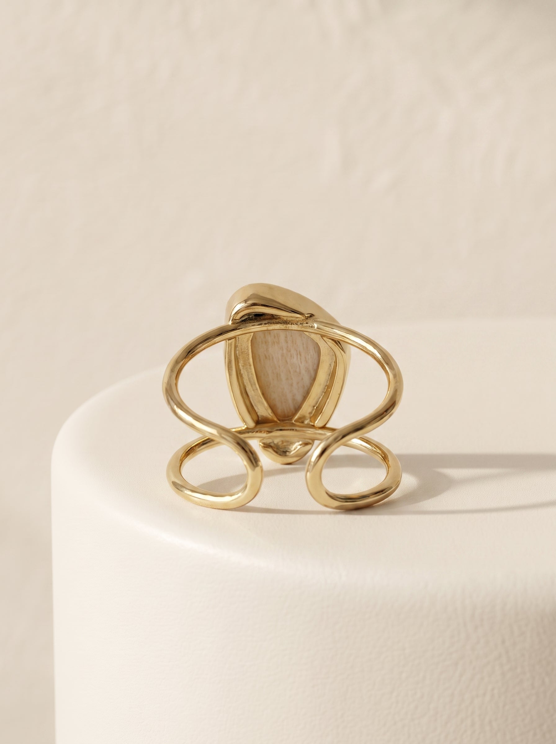 A gold-toned ring with an abstract, looping openwork design and a central oval inset featuring a light beige woodgrain pattern, displayed on a white cylindrical pedestal against a textured cream background.
