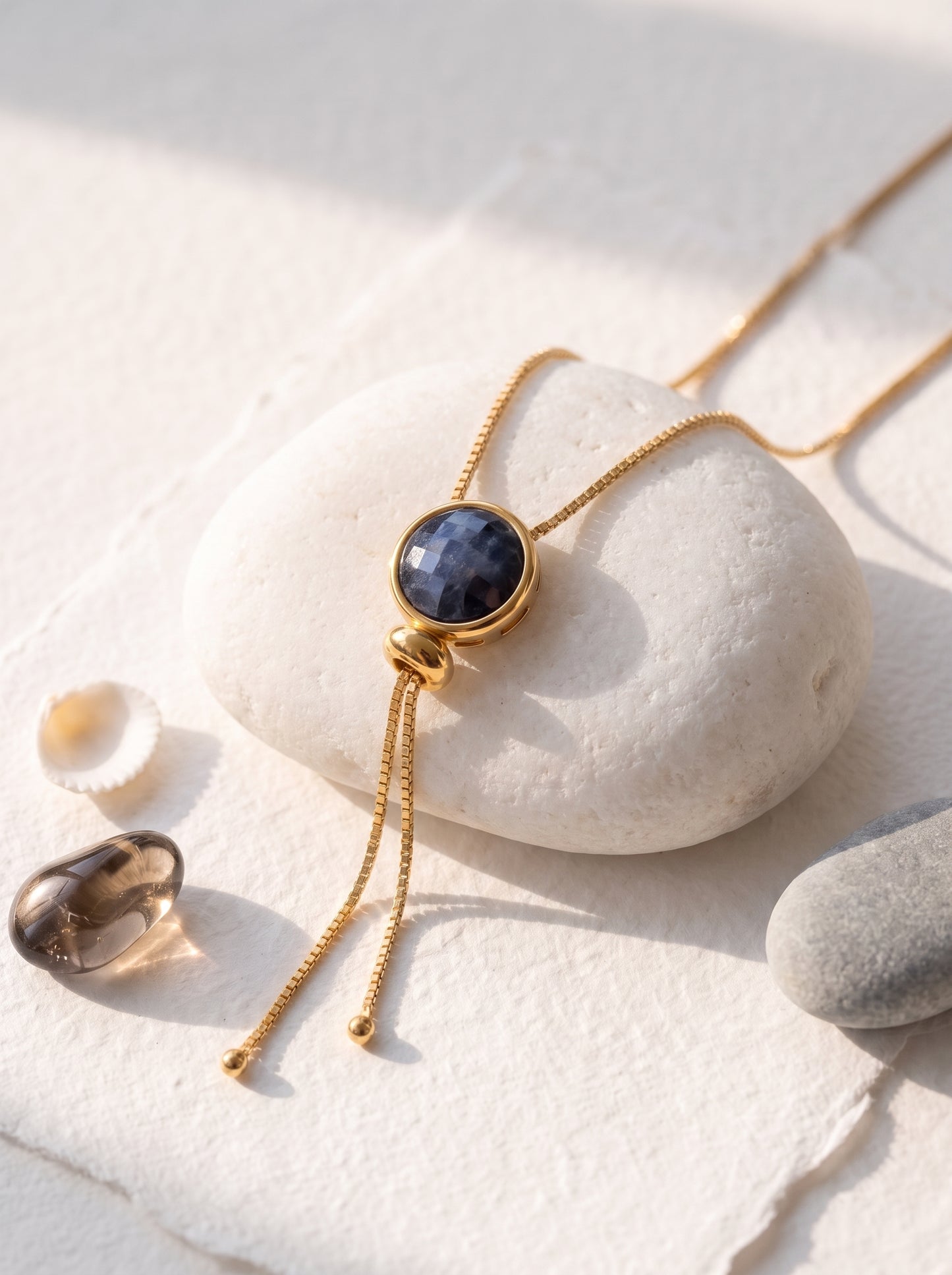 A gold-toned necklace with a round dark blue quartz gemstone pendant and two dangling chains, styled on a white stone with small smooth stones nearby on a textured white surface.