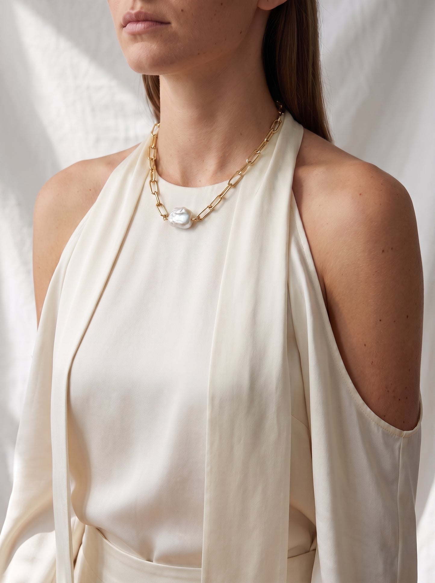 A close-up of a model wearing a gold chain necklace with a large white pearl pendant, styled with a cream halter top against a draped white fabric background.