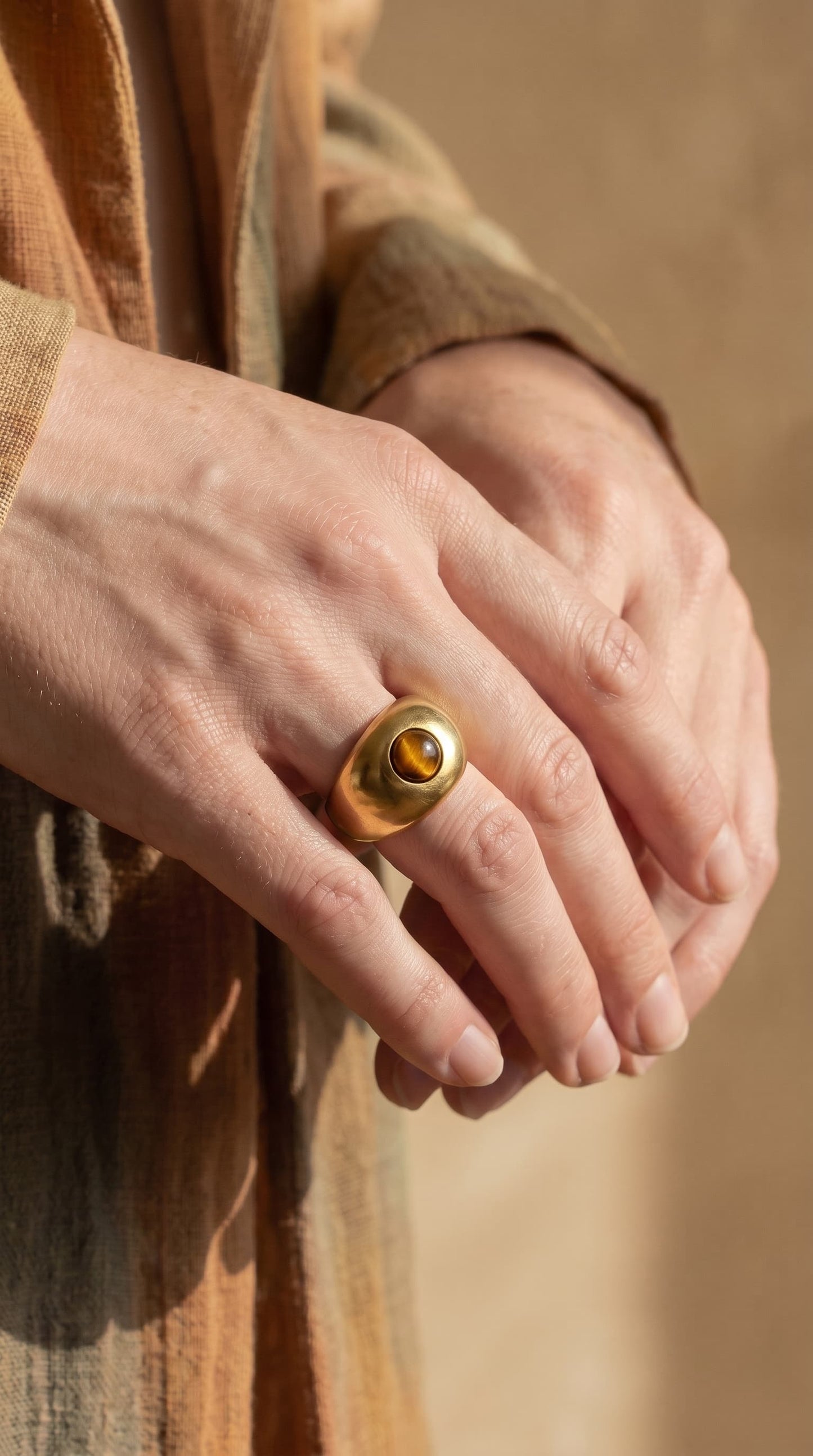 Close-up of model wearing gold-plated tiger's eye dome signet ring on index finger, hand raised near face, wearing a rust-coloured top