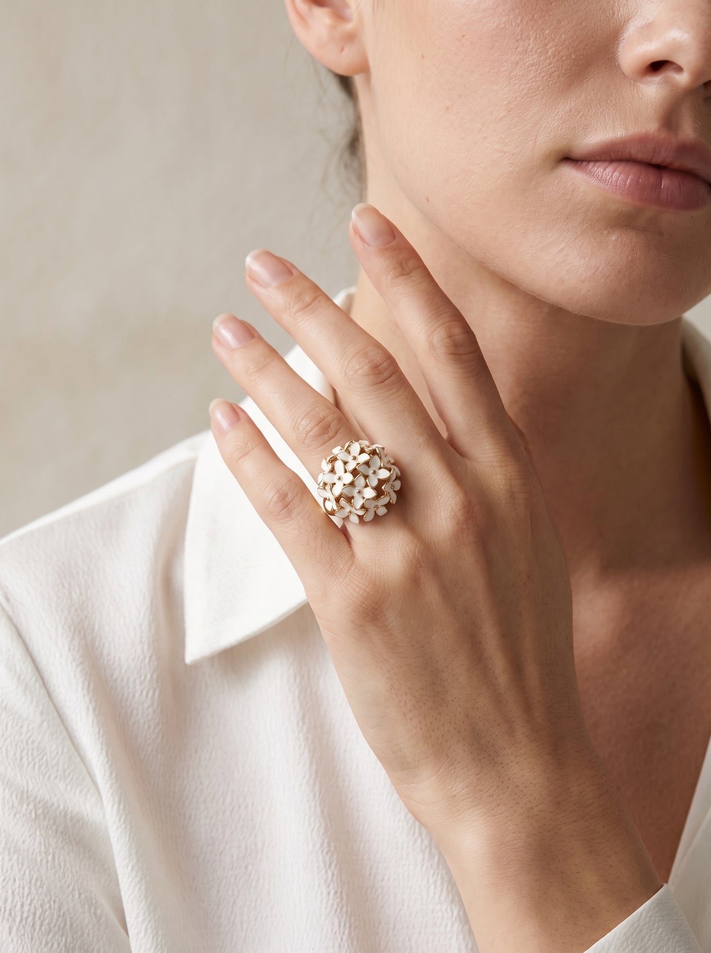 A close-up lifestyle shot of a model wearing a large, textured gold ring with small white floral details on her finger, paired with a white collared blouse against a soft beige background.