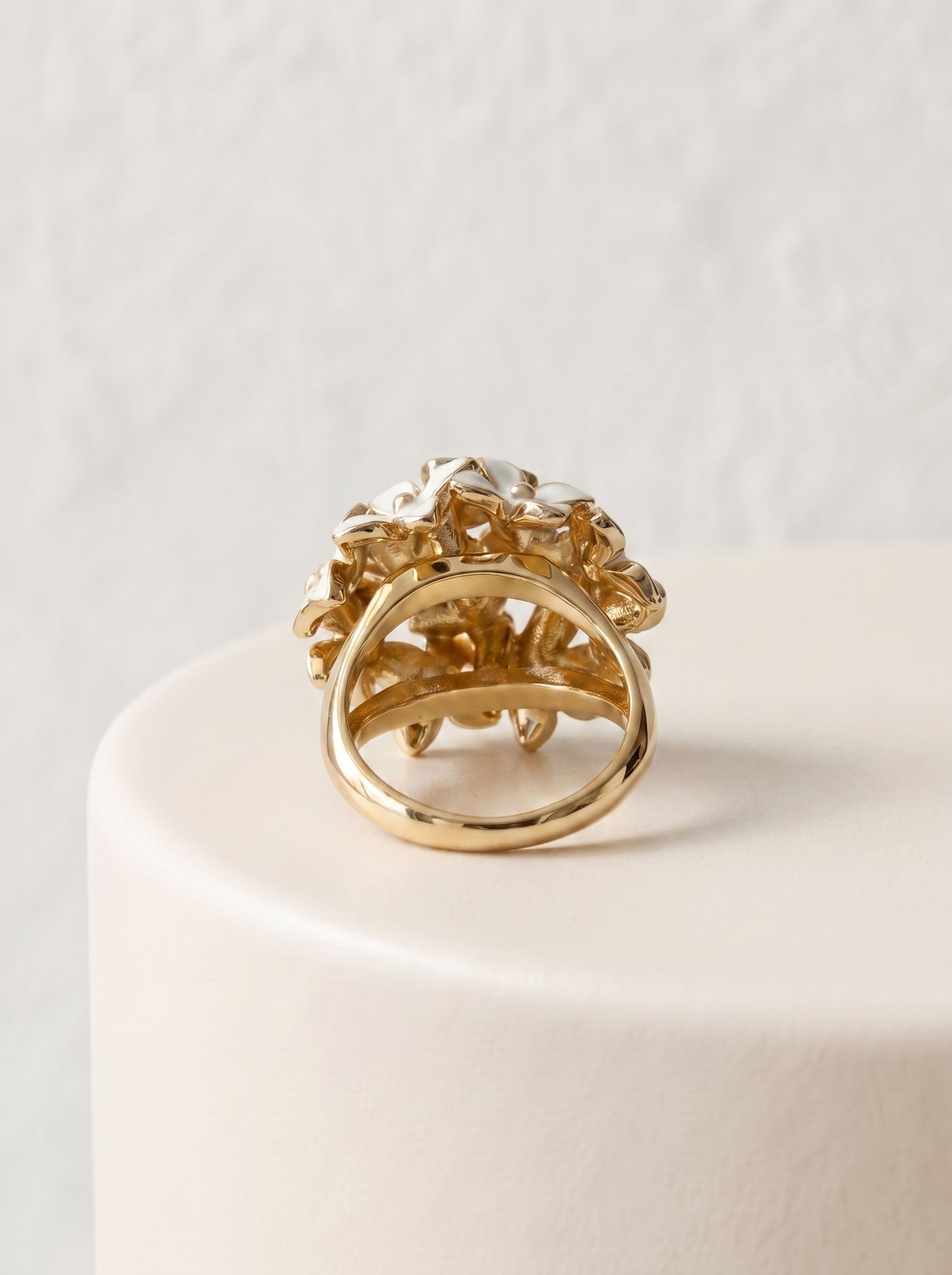 A gold ring with a textured, sculptural floral or leaf-like top and a smooth circular band, displayed on a white cylindrical pedestal against a light textured wall.