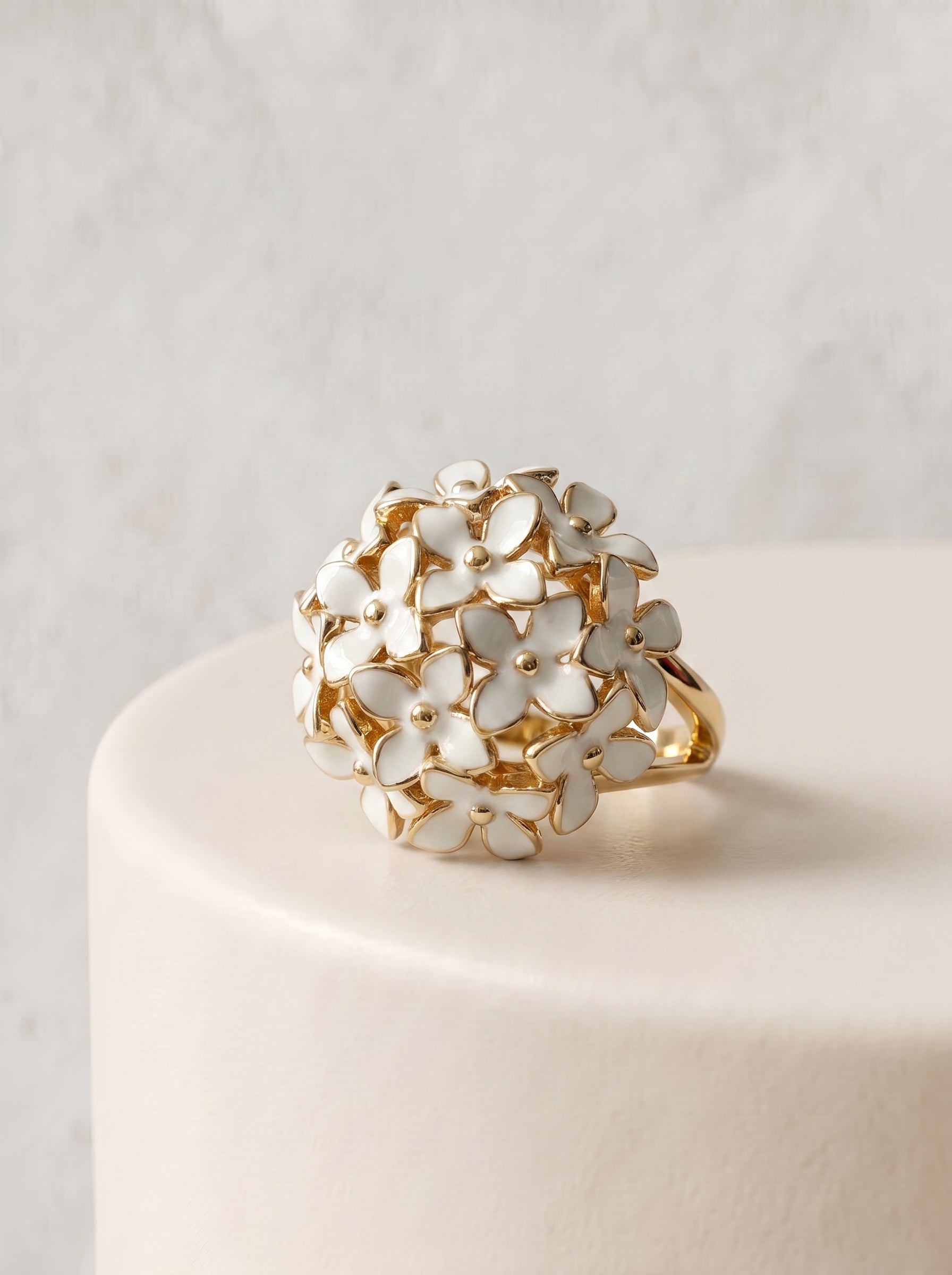 Gold-plated dome ring with white enamel flower cluster displayed on a white cylindrical stand