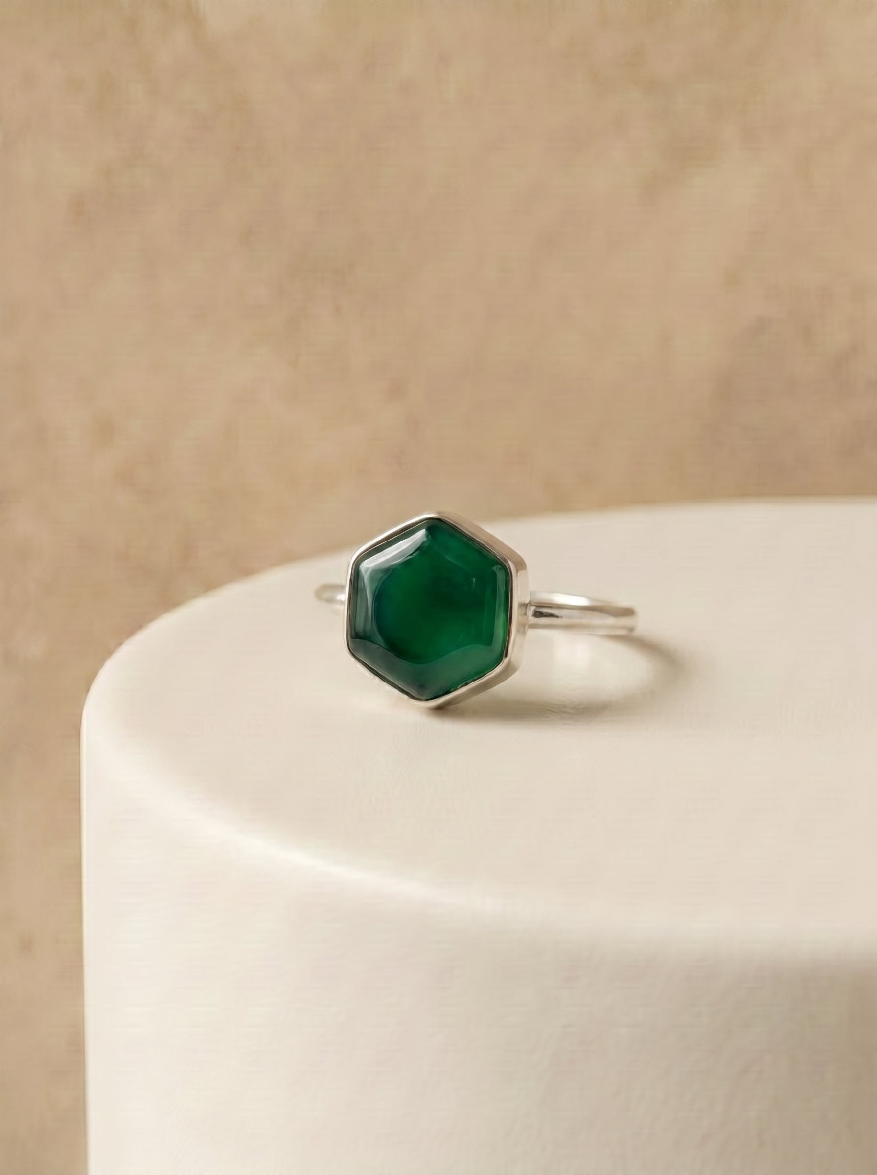 A silver ring with a faceted green  onyx gemstone, displayed on a white cylindrical pedestal against a textured beige background.