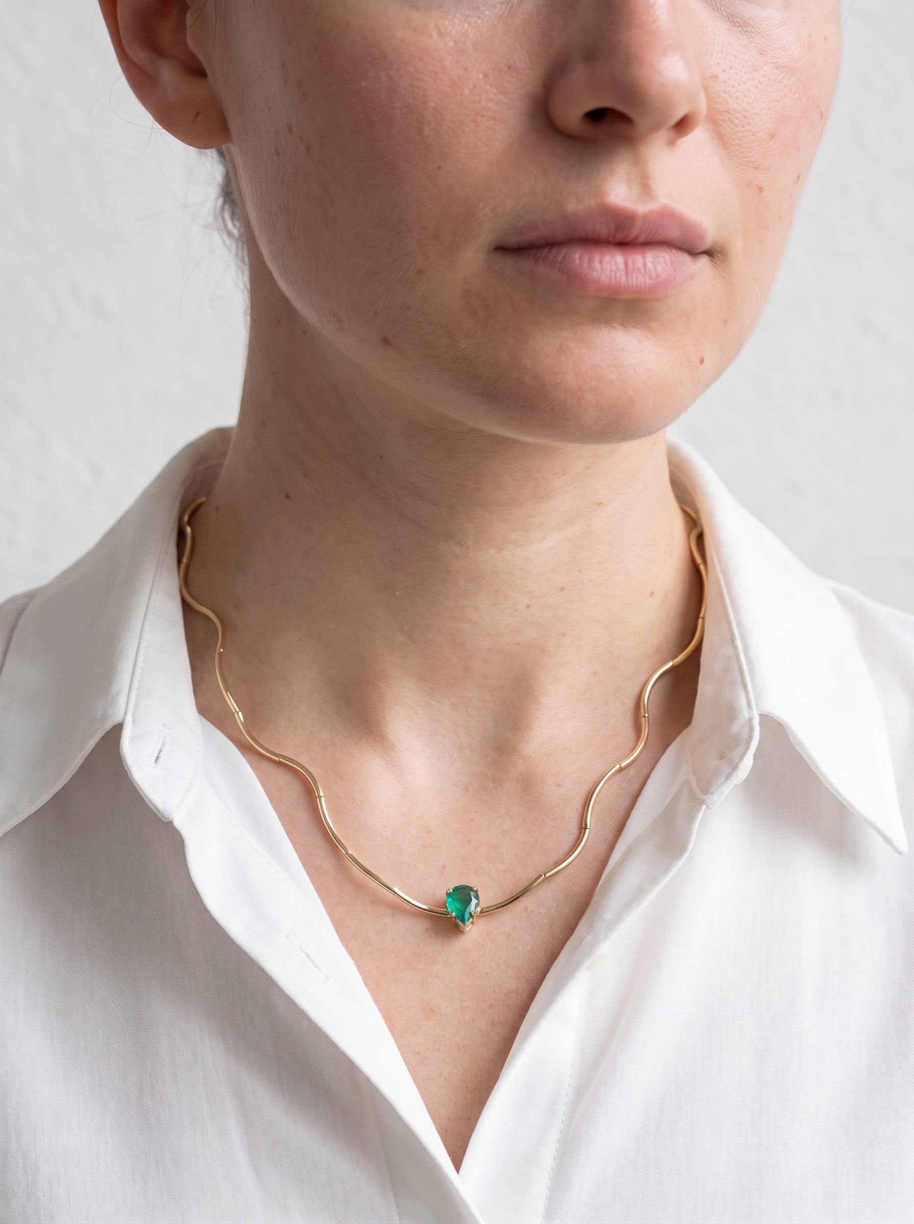 Gold-plated bamboo bar chain necklace with green crystal solitaire worn by a woman in an open-collar white shirt