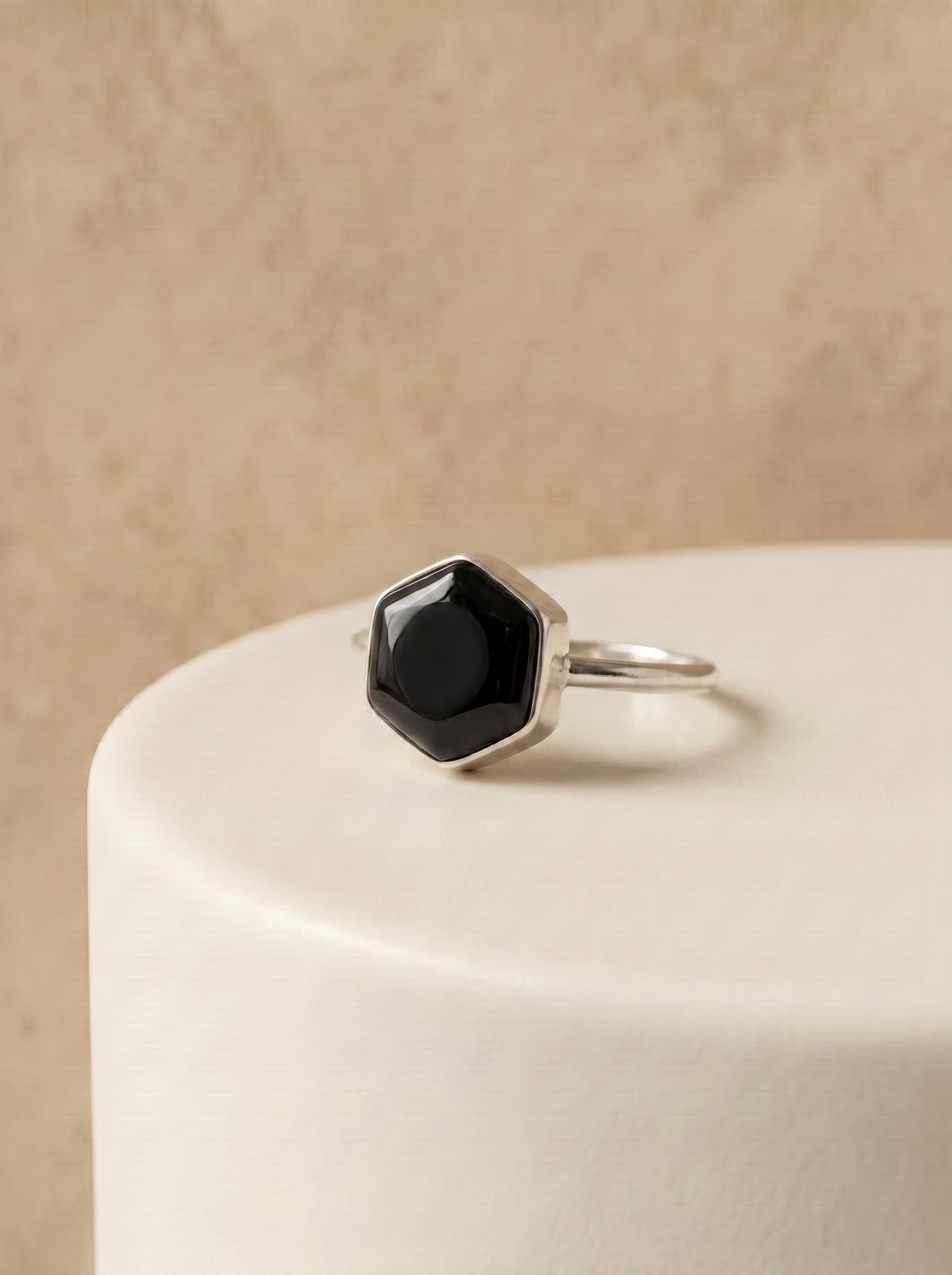A silver ring with a faceted black onyx stone, displayed on a white cylindrical pedestal against a textured beige background.