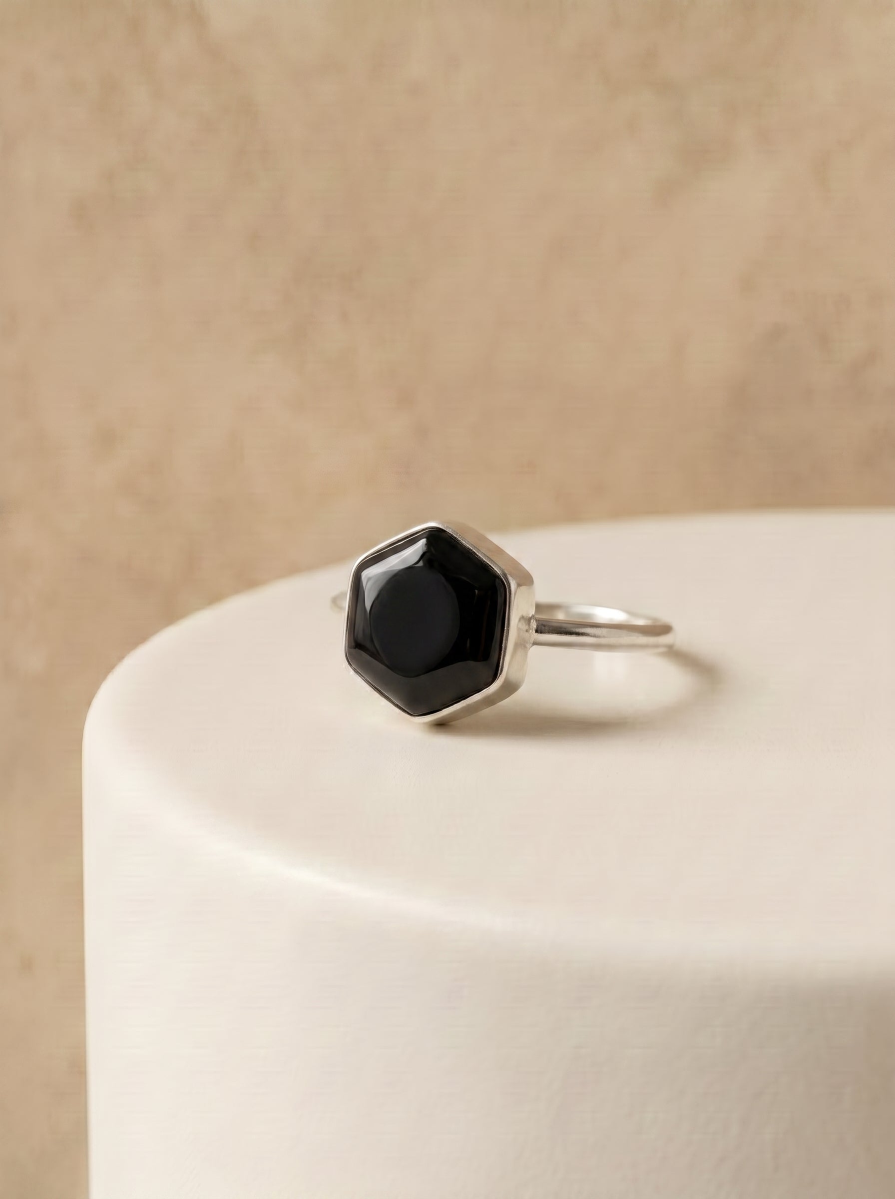 A silver ring with a faceted black onyx stone, displayed on a white cylindrical pedestal against a textured beige background.