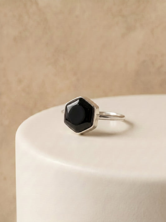 A silver ring with a faceted black onyx stone, displayed on a white cylindrical pedestal against a textured beige background.