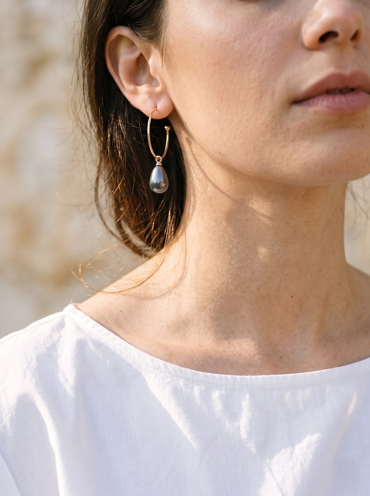 Close-up model: “Close-up of a woman wearing a StoneCraft Jewellery grey pearl drop earring on a gold-plated open hoop.
