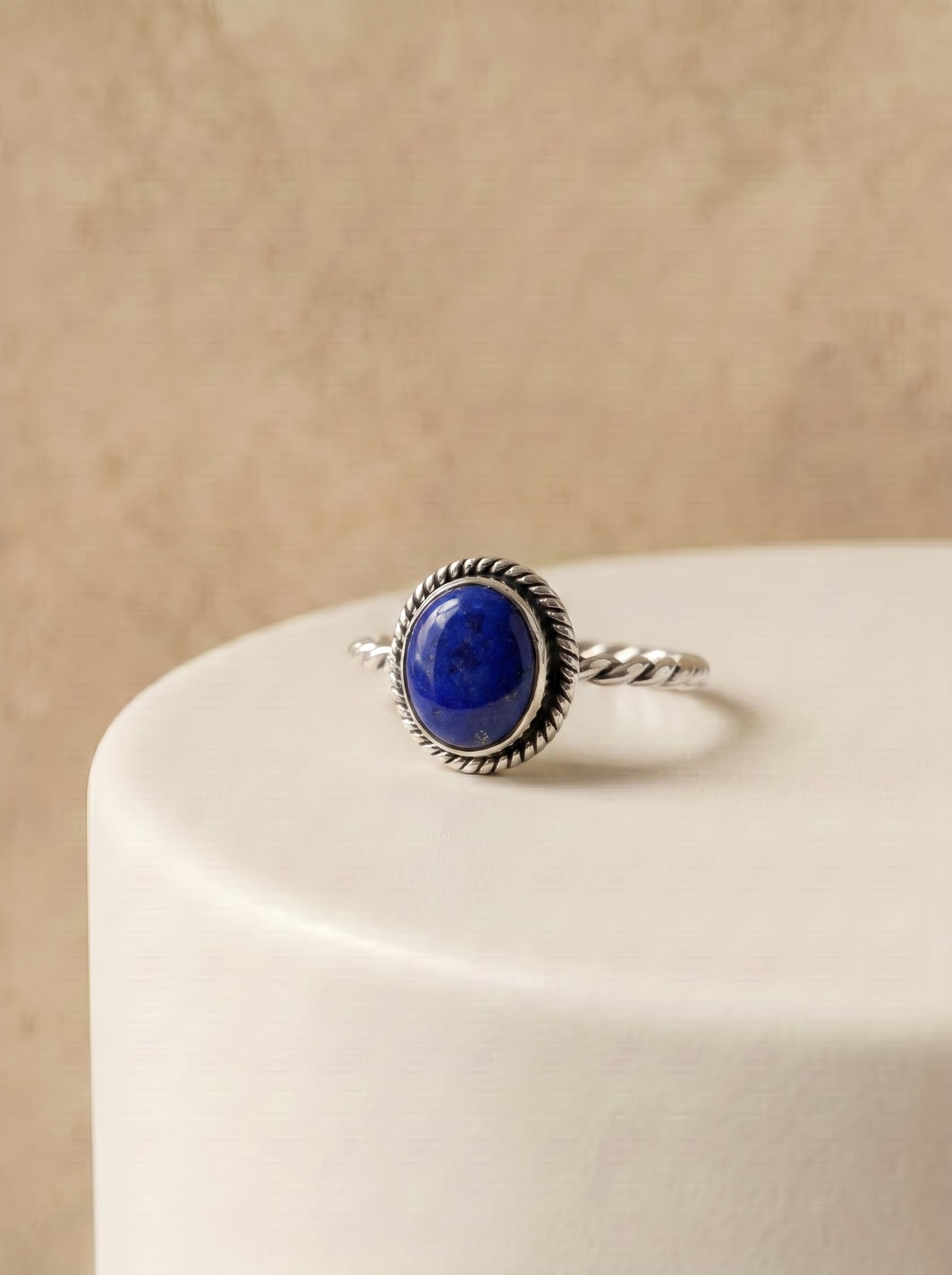 Sterling silver boho ring with oval cabochon lapis lazuli gemstone and twisted rope-detail band, displayed on a white ceramic stand

