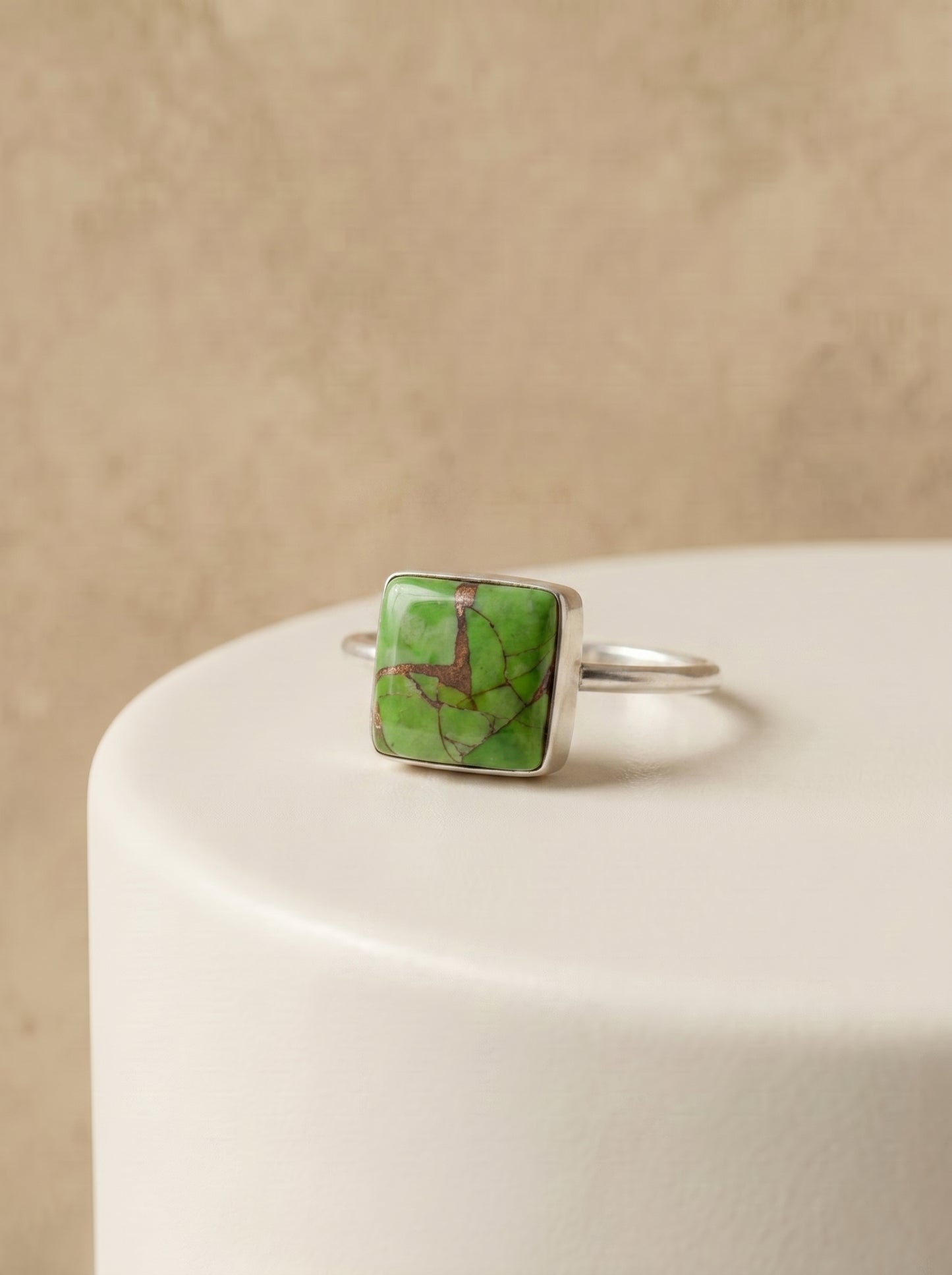 A silver ring with a square green stone featuring natural brown veining, displayed on a white cylindrical pedestal against a textured beige background.