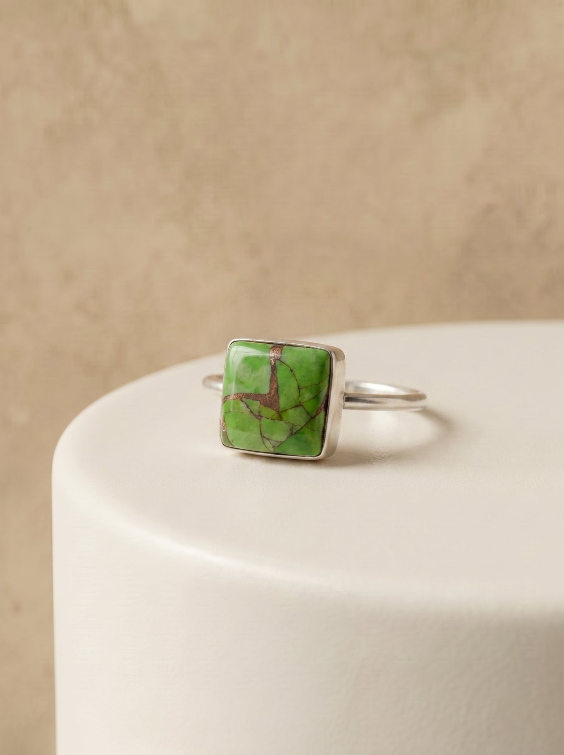 A silver ring with a square green stone featuring natural brown veining, displayed on a white cylindrical pedestal against a textured beige background.