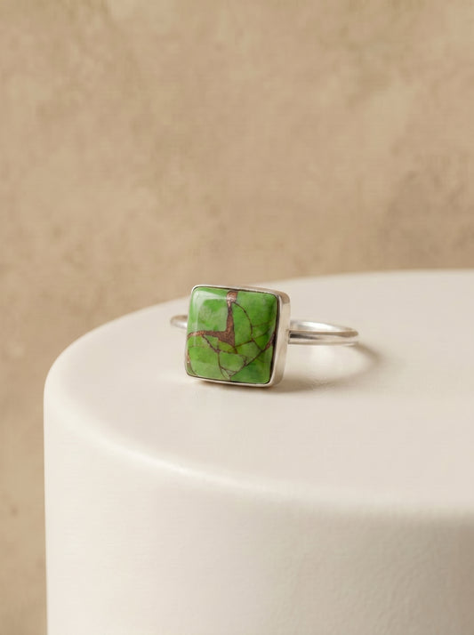 A silver ring with a square green stone featuring natural brown veining, displayed on a white cylindrical pedestal against a textured beige background.