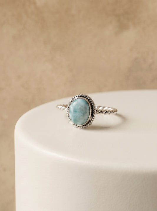 A silver ring with a round larimar stone and a textured rope border, displayed on a white cylindrical pedestal against a warm beige background.