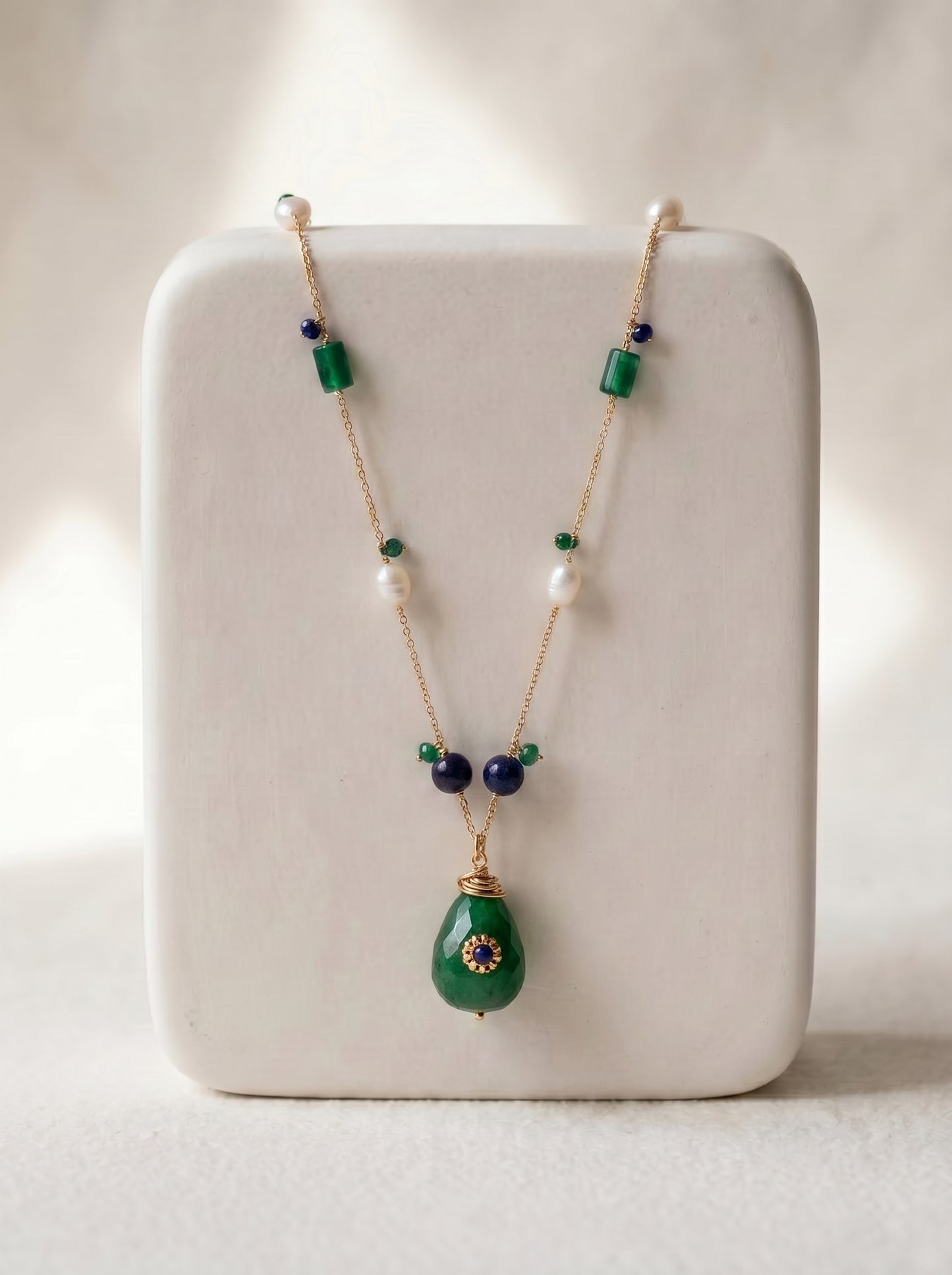 Gold-plated long necklace with green jade teardrop pendant, sapphire floral cap, freshwater pearls and jade cylinder beads on a delicate chain, displayed on a white ceramic jewellery stand