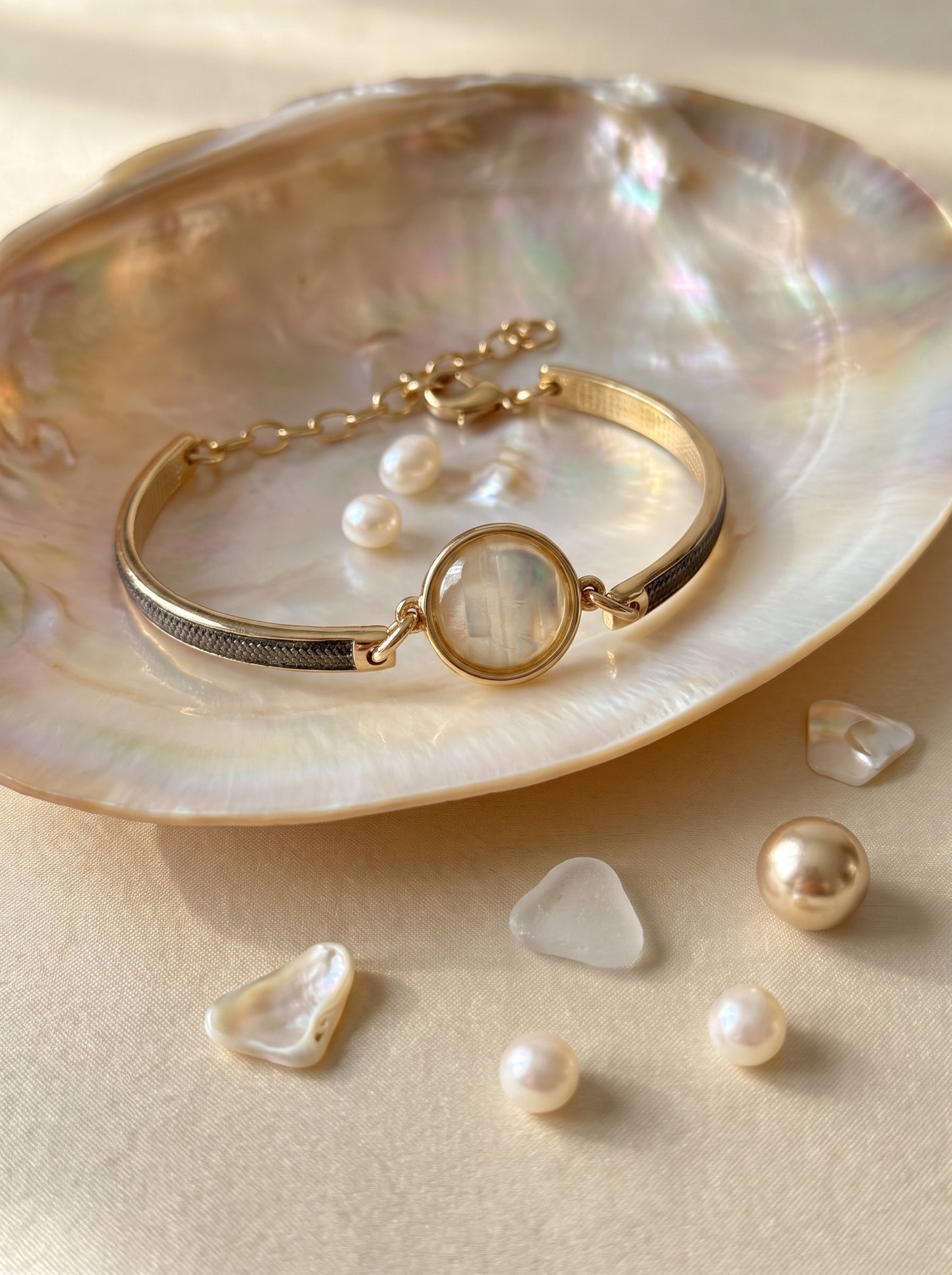 A gold-toned  plated and rhodium plated bangle bracelet with a round iridescent nautilus centerpiece, displayed on a large iridescent shell with small pearls scattered around on a warm beige surface.