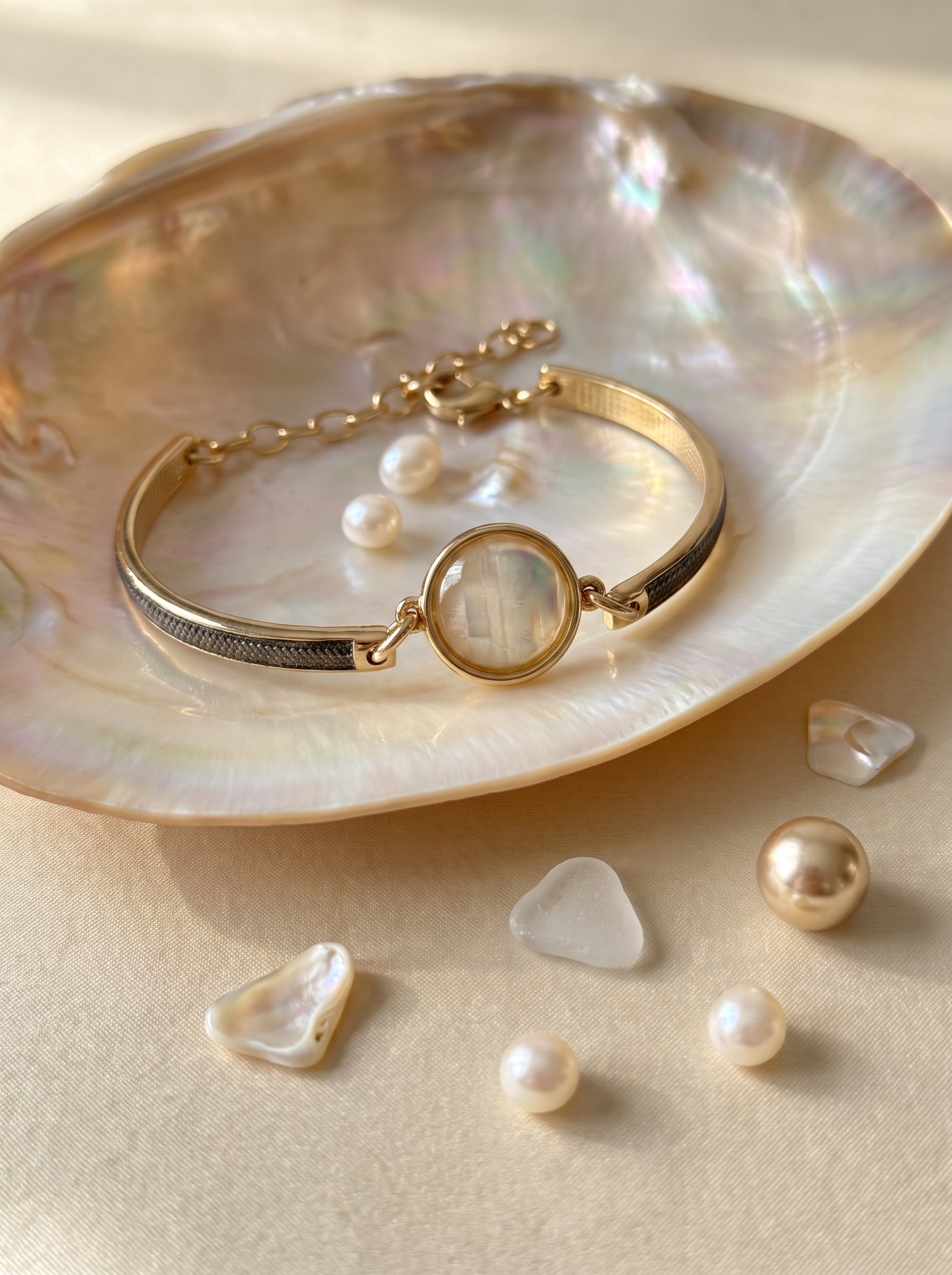 A gold-toned  plated and rhodium plated bangle bracelet with a round iridescent nautilus centerpiece, displayed on a large iridescent shell with small pearls scattered around on a warm beige surface.